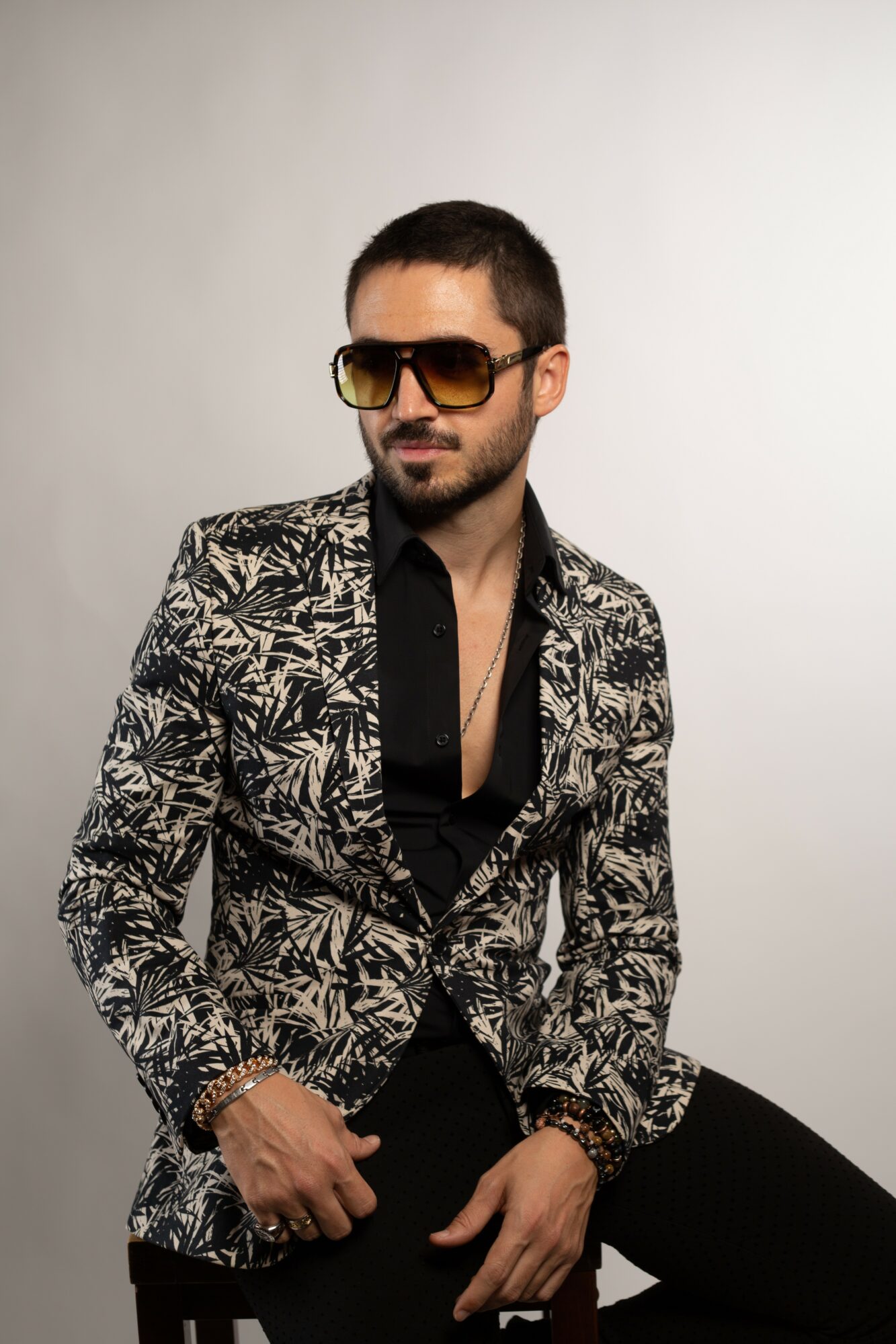 Man with short dark hair, beard, sunglasses, wearing a patterned blazer and black shirt, sitting against a plain background.