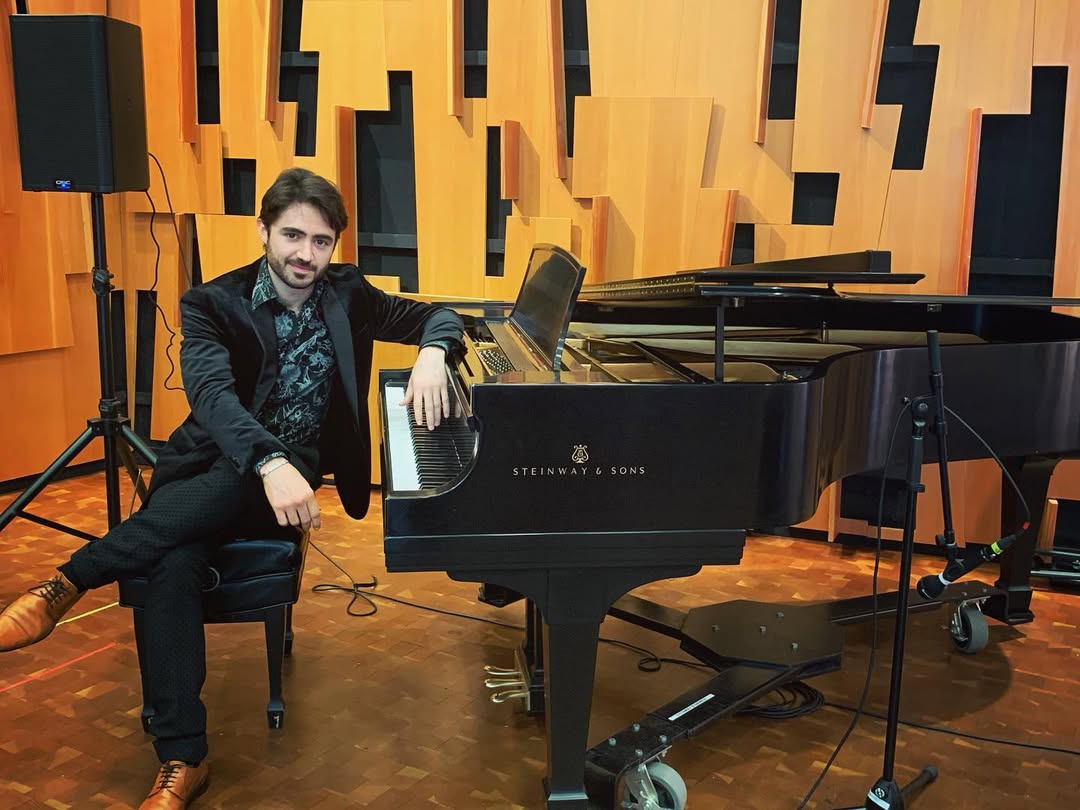 Man sitting on a chair next to a grand piano in a music room with wooden wall panels.