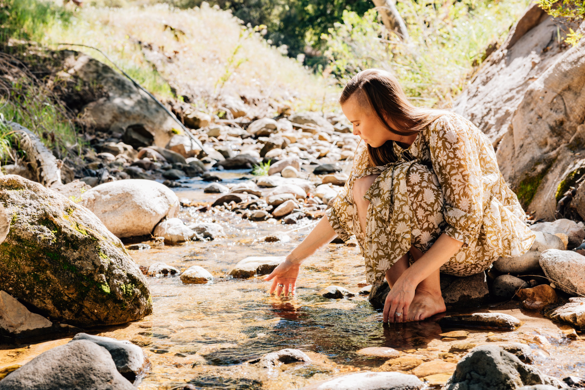 Woman crouching by a rocky stream, reaching into the water outdoors during daytime.