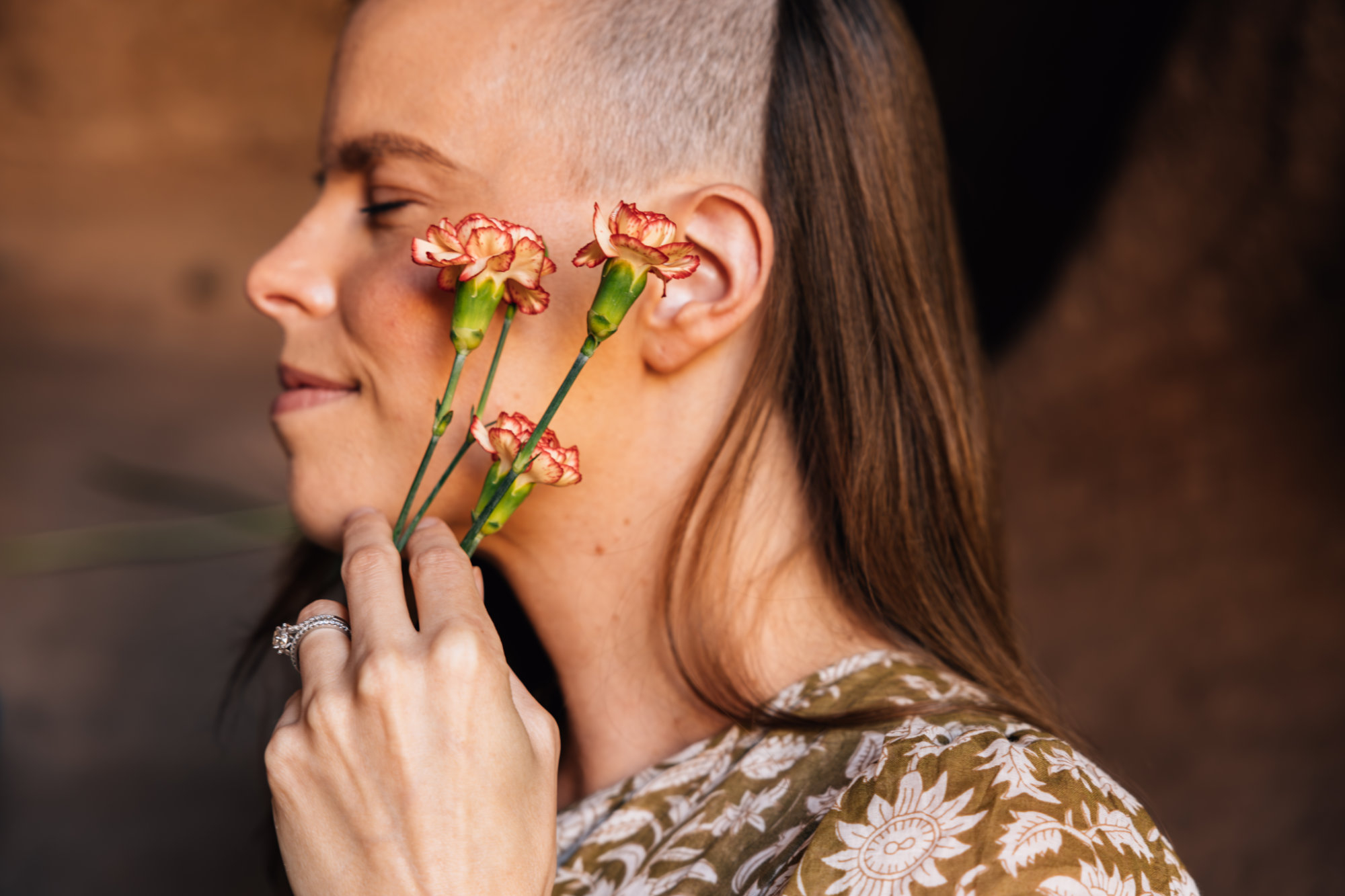 Woman with a shaved side hairstyle holding pink flowers near her face, smiling gently, wearing a patterned top.