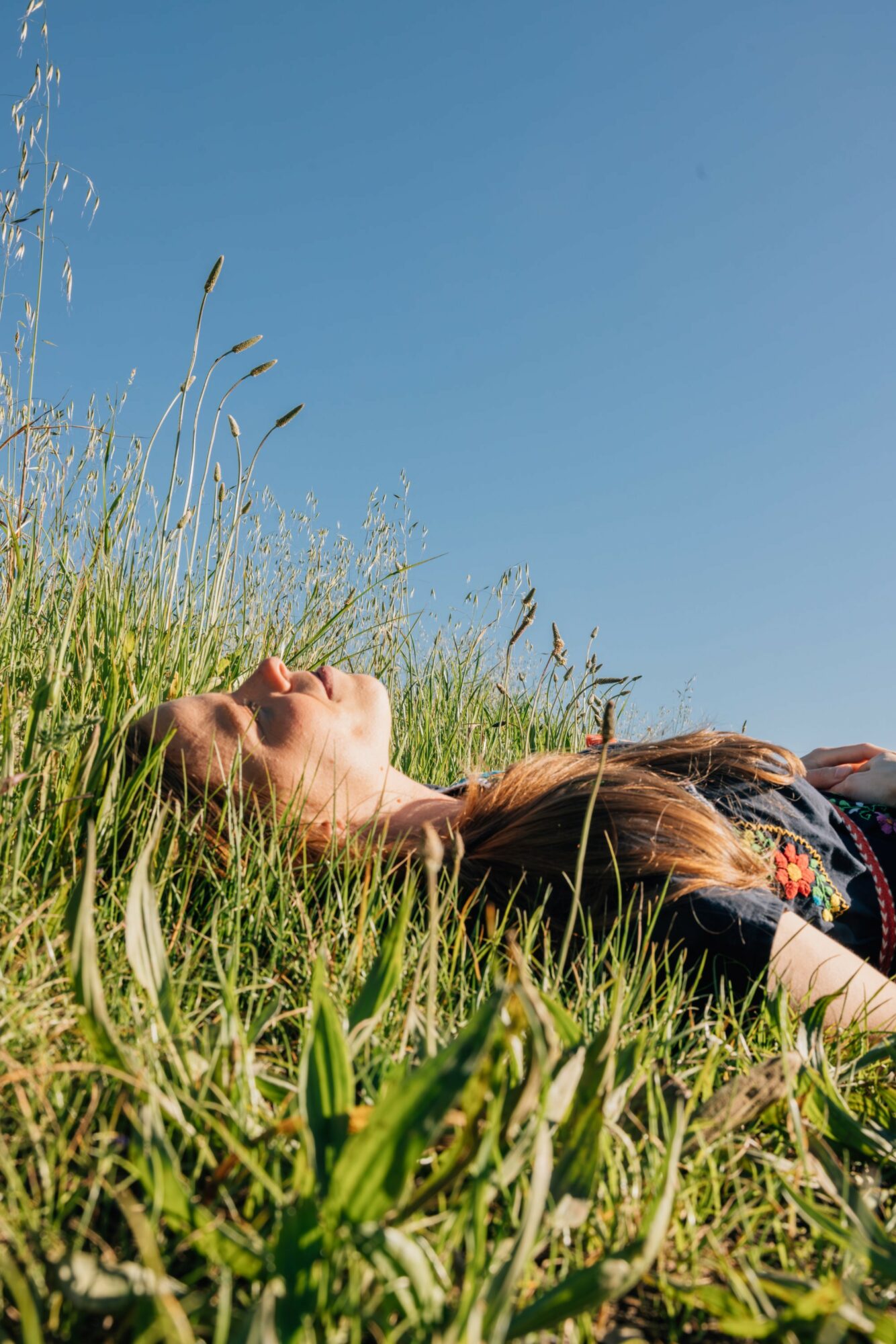 Person lying on grass with arms outstretched, head tilted back, under a clear blue sky.
