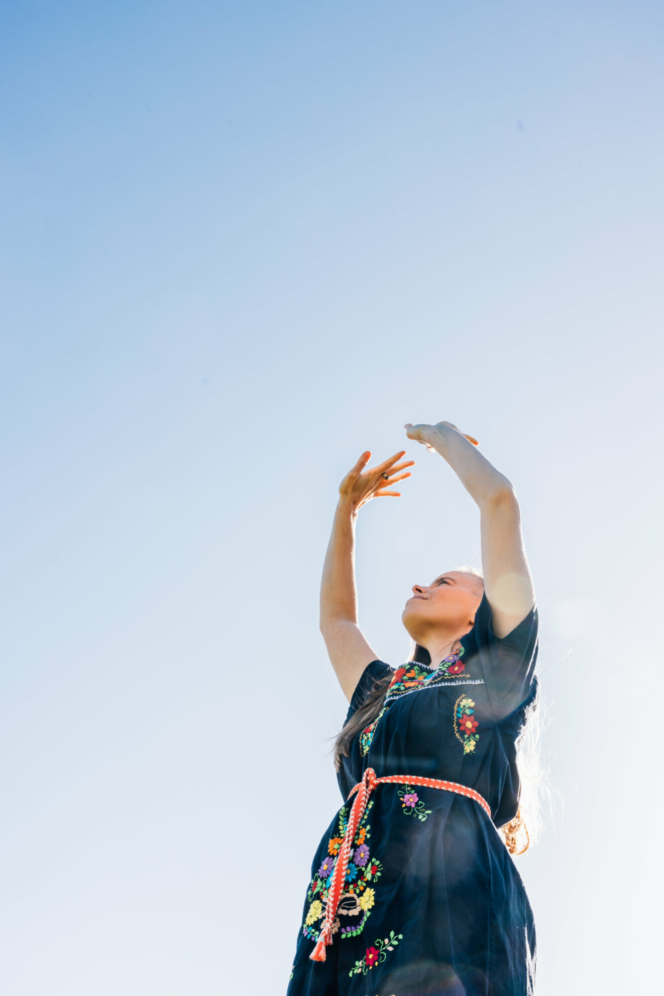 Person in a dark dress with floral patterns, arms raised, looking upward against a clear sky.