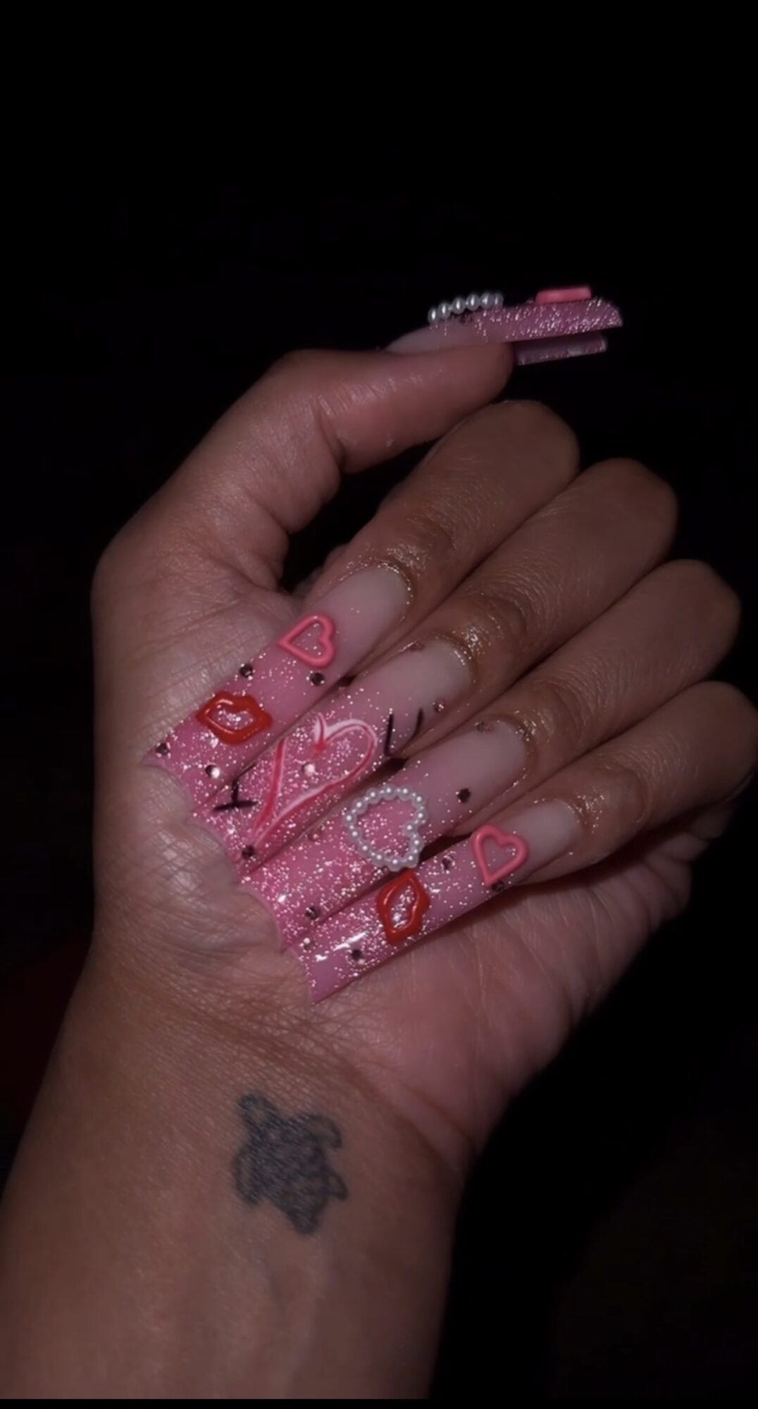 Hand with decorated nails featuring pink, red, and white designs, and a small tattoo of a bear on the wrist.