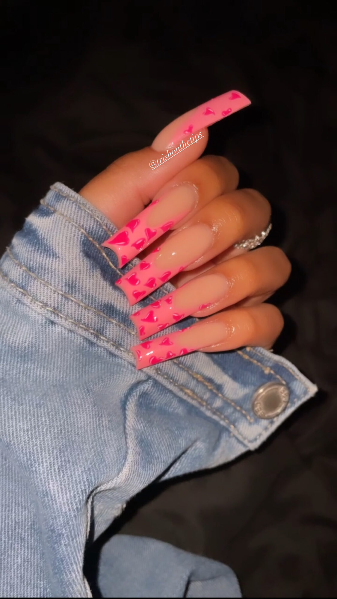 Hand with long pink and clear acrylic nails, wearing a denim jacket, against a dark background.