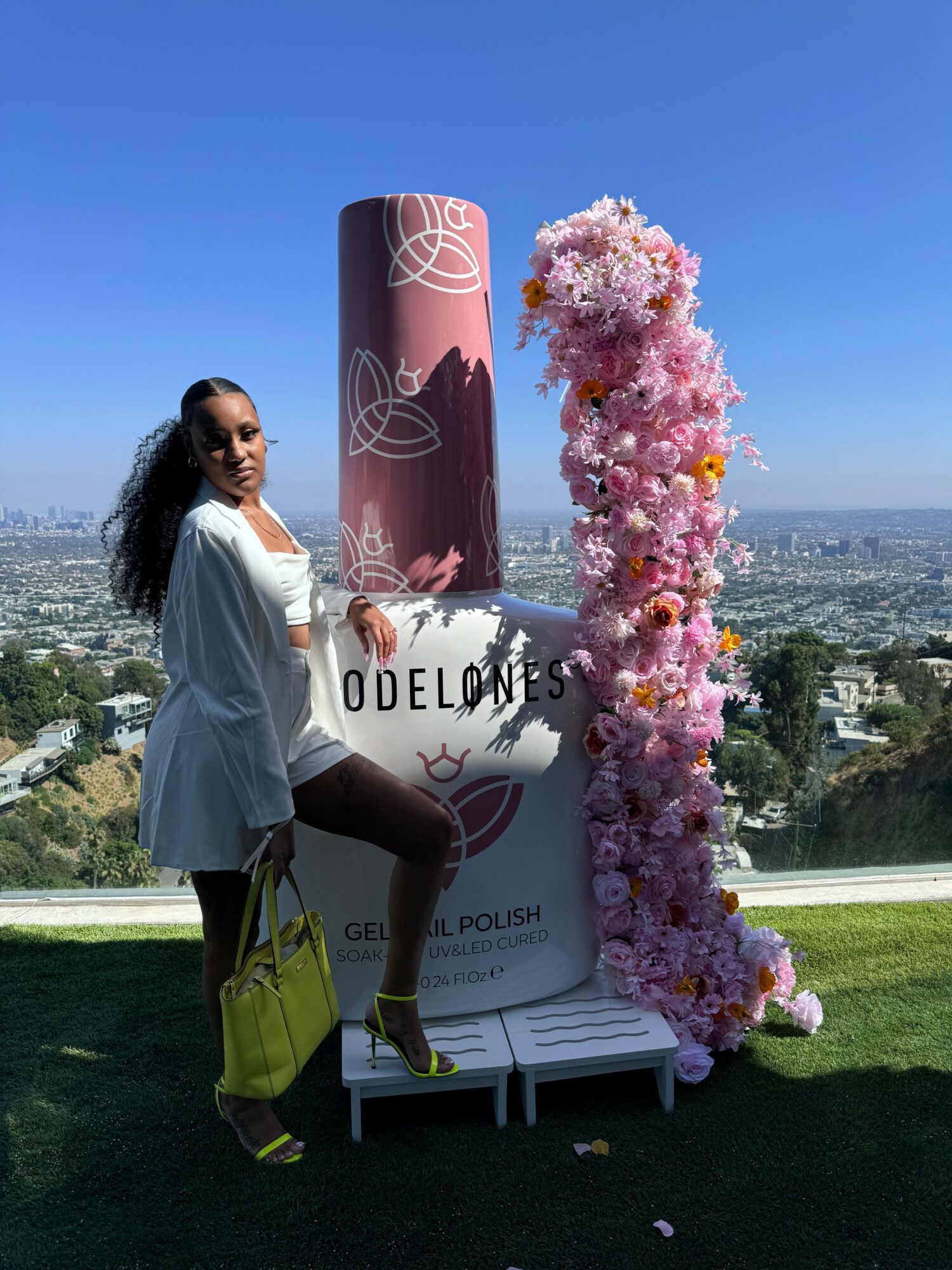 Woman standing outdoors next to a large pink and white floral display and a cylindrical sign with text, cityscape in background.