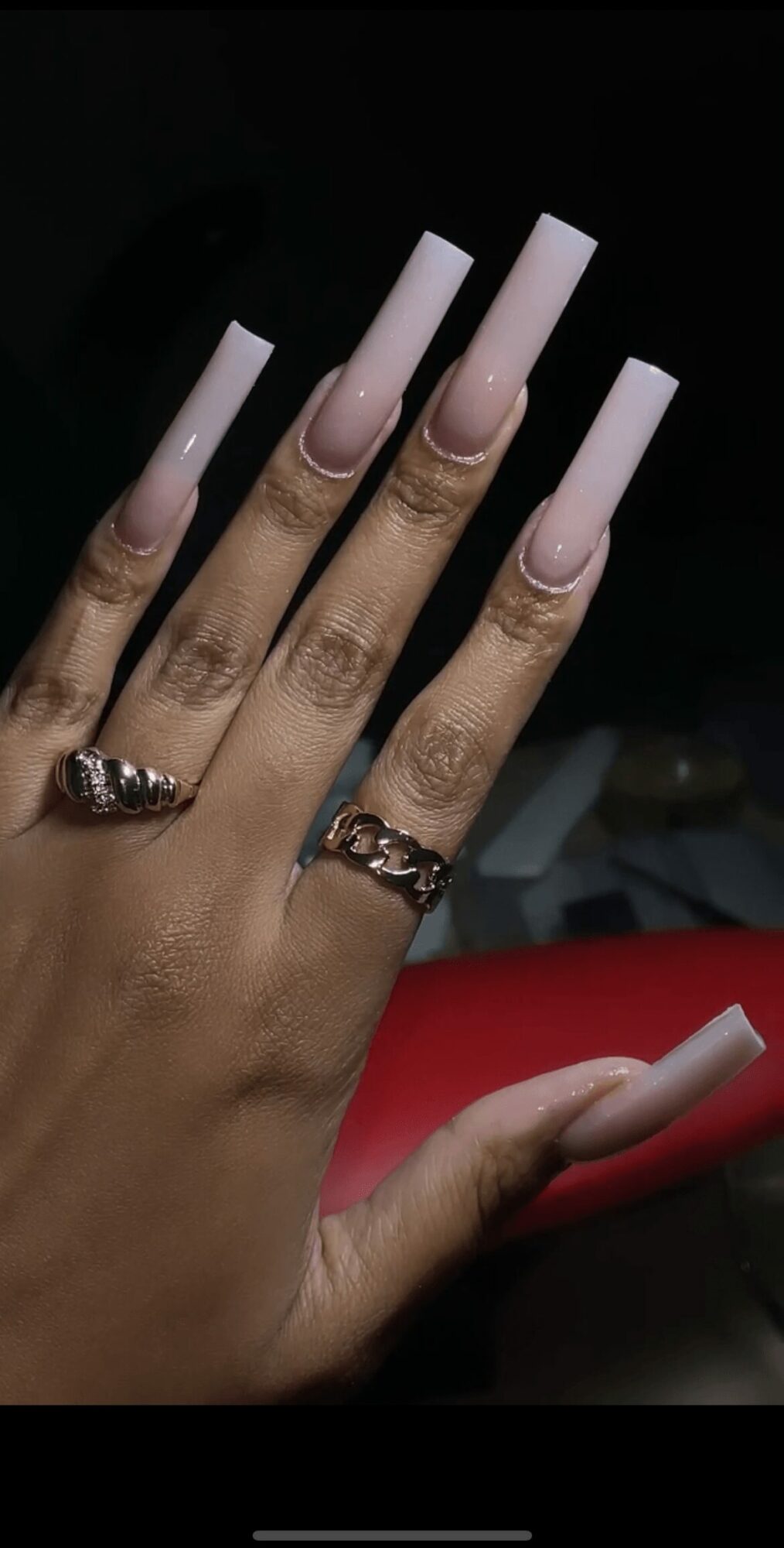 Hand with long, square-shaped, light pink nails and rings on fingers, against a dark background.