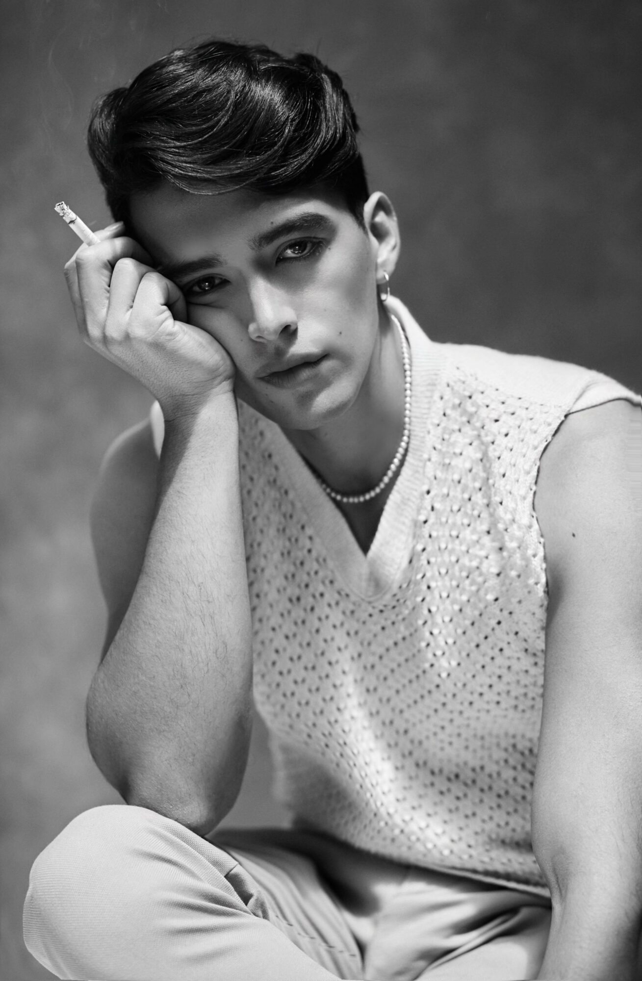 Young person with short hair, resting face on hand, holding cigarette, wearing sleeveless top, in black and white.