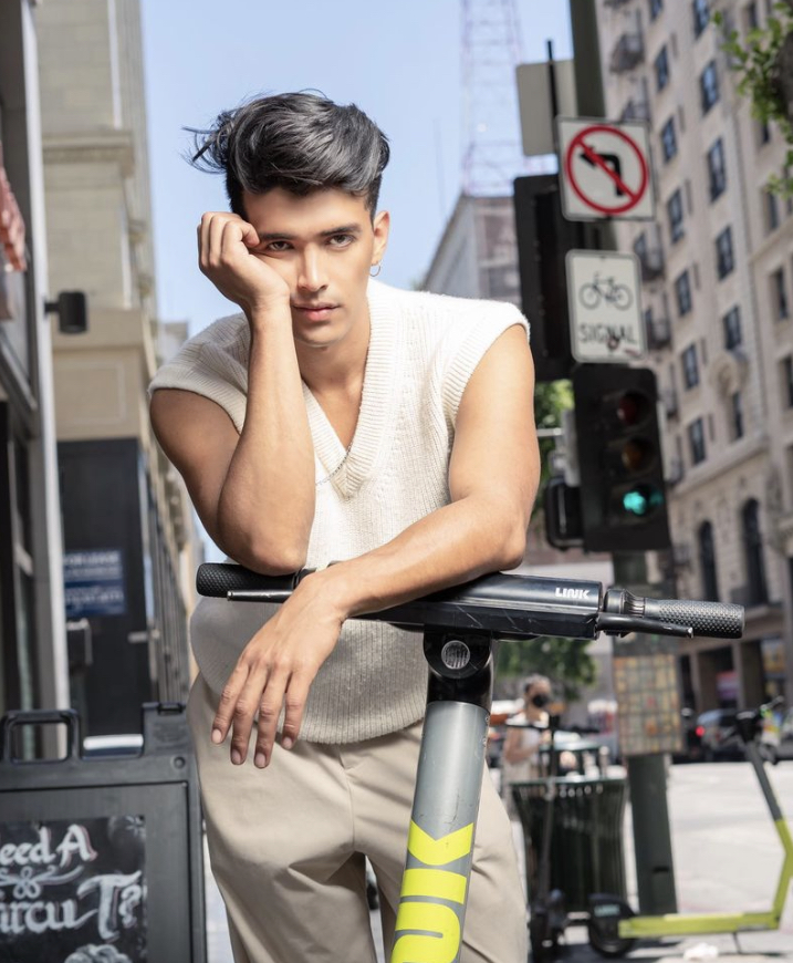 Young man leaning on a scooter on a city street with buildings and traffic signals in background.