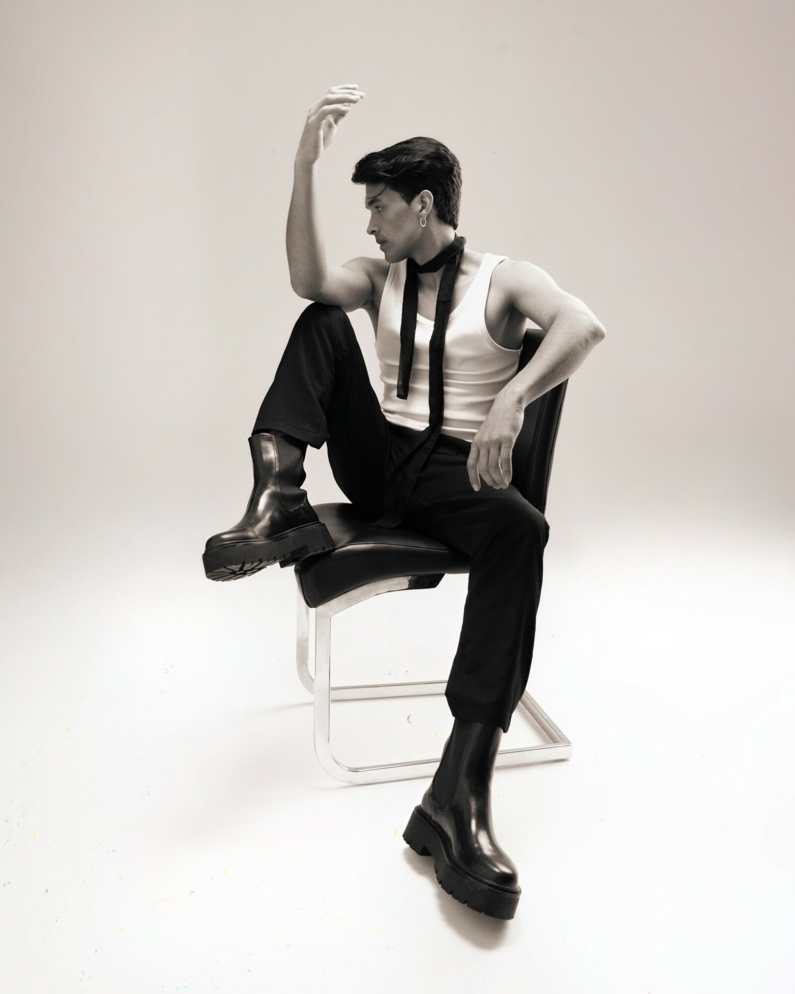 Person sitting on a chair with one leg raised, wearing boots, suspenders, and a sleeveless top, in a neutral setting.