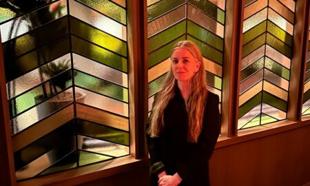 A woman with long hair stands indoors in front of decorative glass windows with geometric patterns.