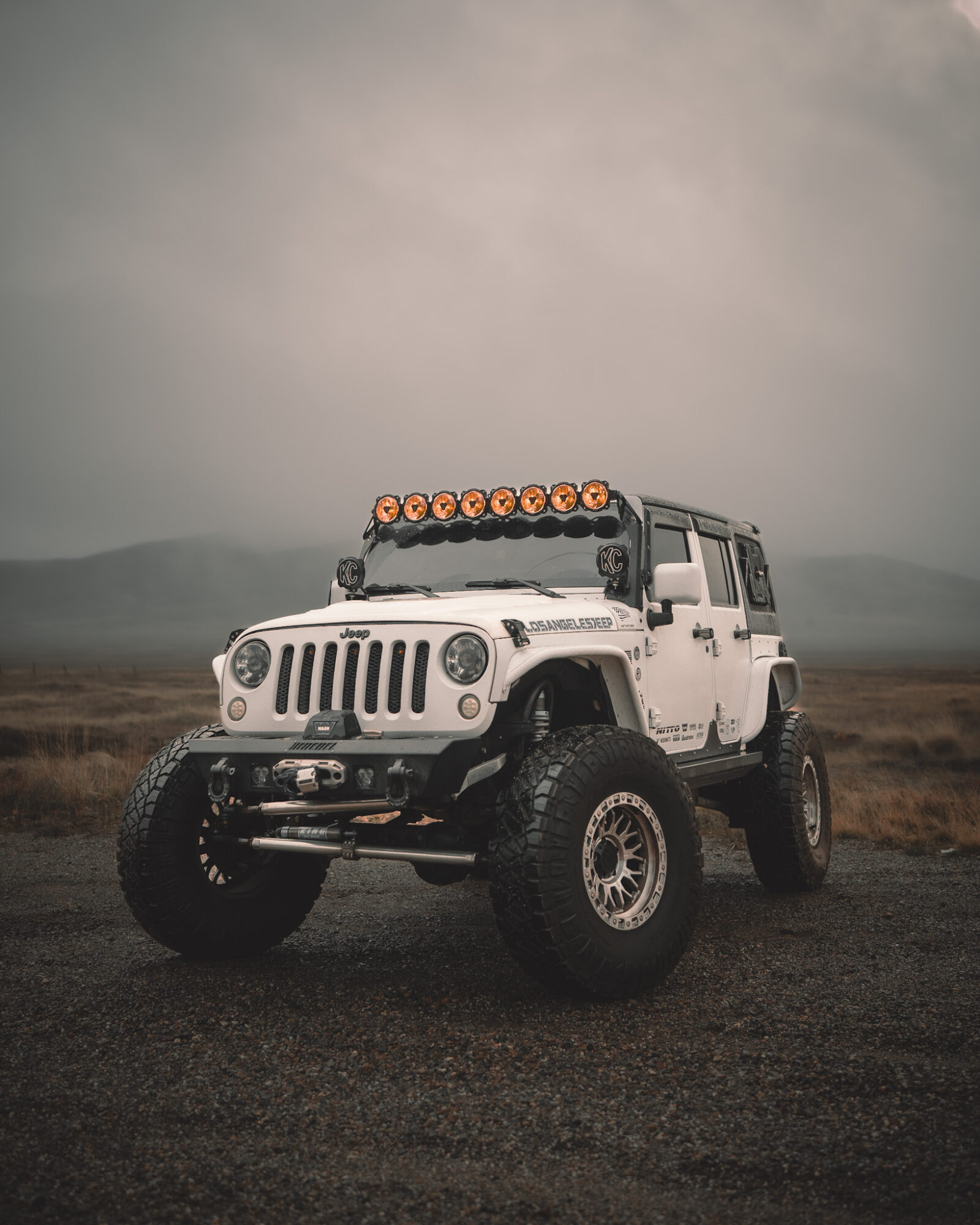 White off-road vehicle with large tires and roof lights parked on a flat landscape under cloudy sky.