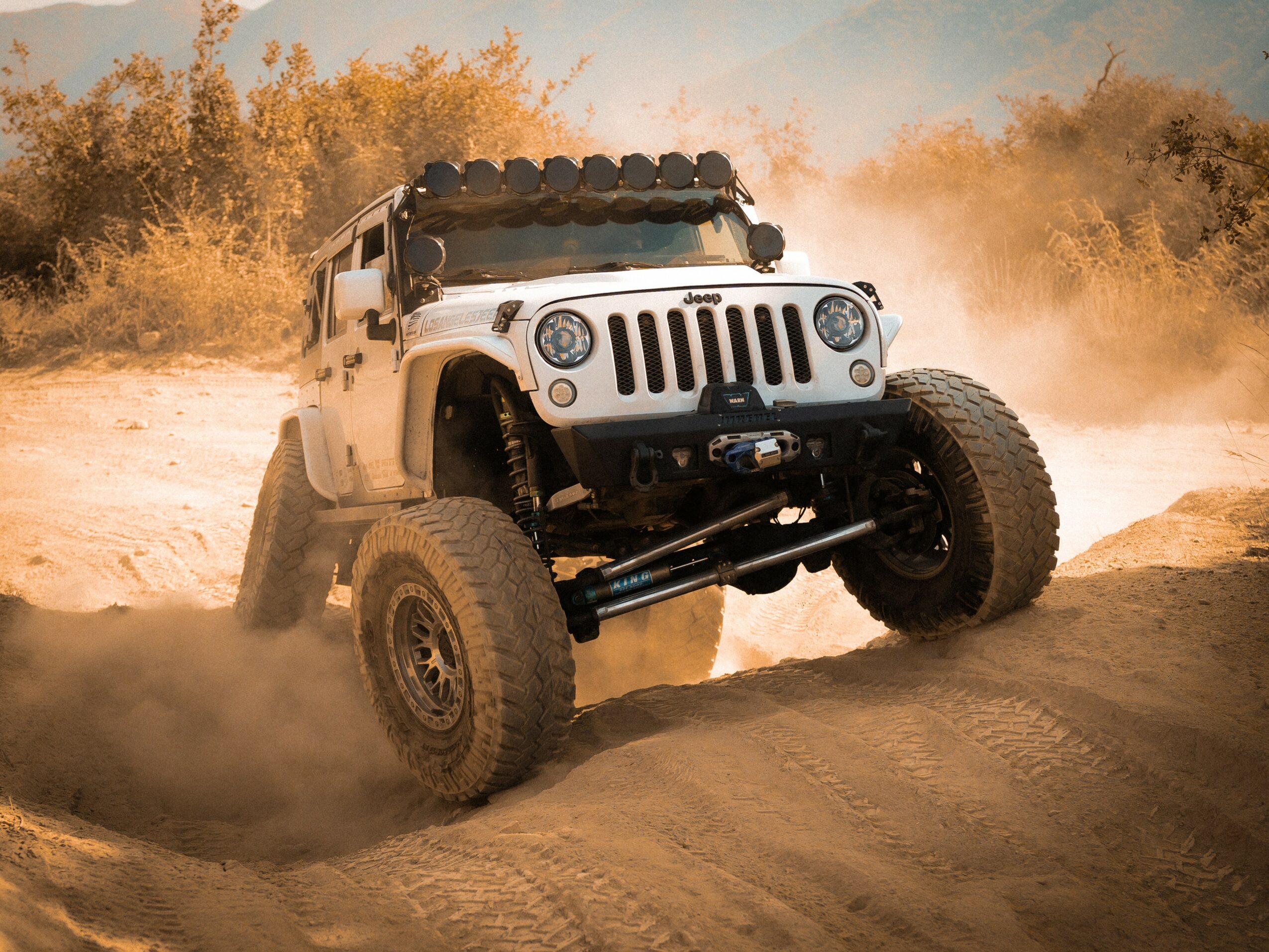 White off-road vehicle driving on sandy terrain with dust and trees in background.