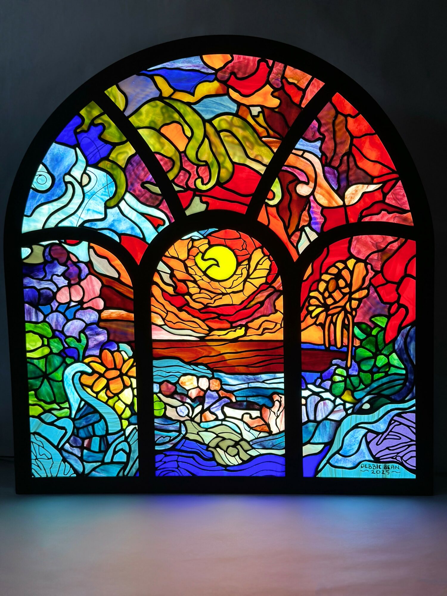 Colorful stained glass window depicting a sunset over a landscape with trees, water, and sky in vibrant colors.
