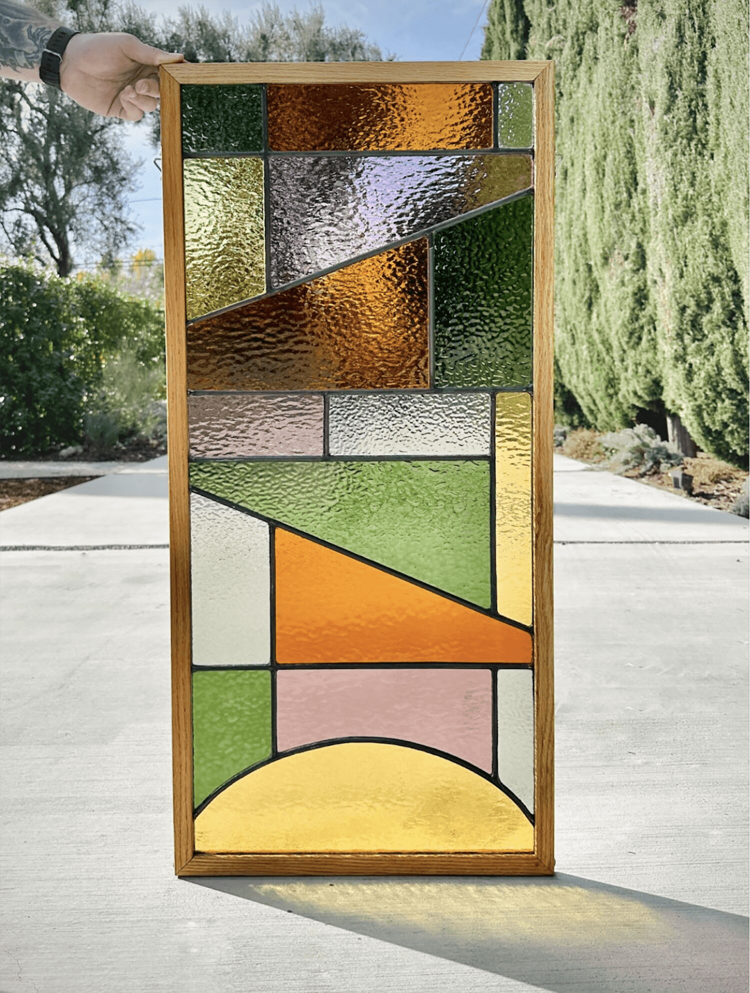 Colorful stained glass window with geometric shapes, held outdoors with trees and sky in background.