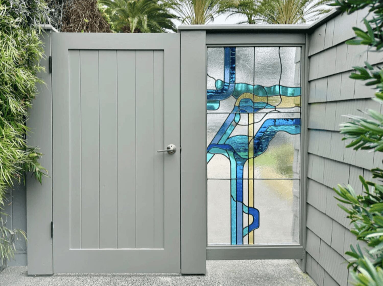 Gray wooden gate with a stained glass window, surrounded by greenery and gray siding.