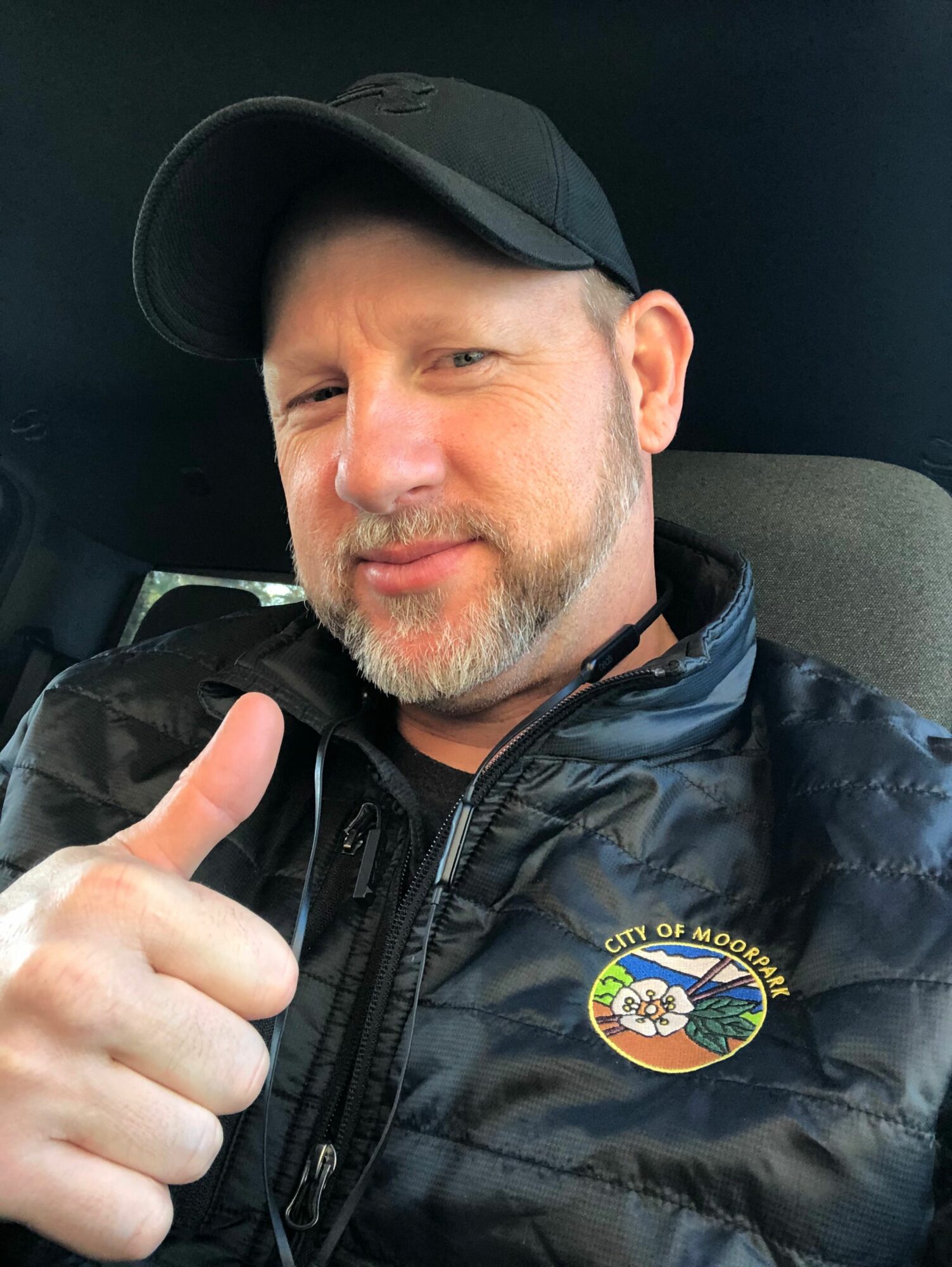 Man wearing a black cap and jacket, giving a thumbs-up, seated indoors with a dark background.