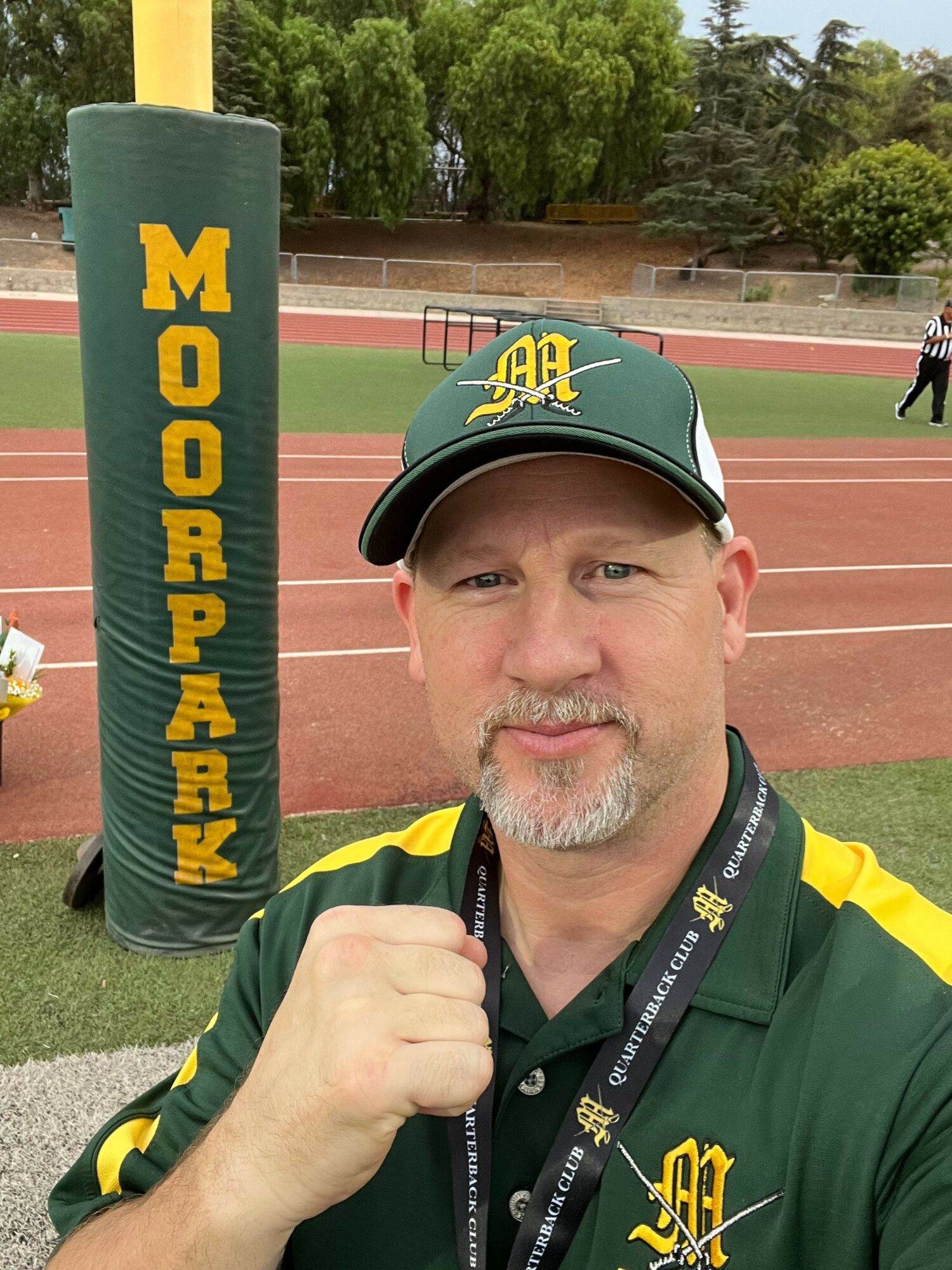 Man in green cap and jacket taking selfie at track field with a pole vault padding that reads 'Moorpark'.