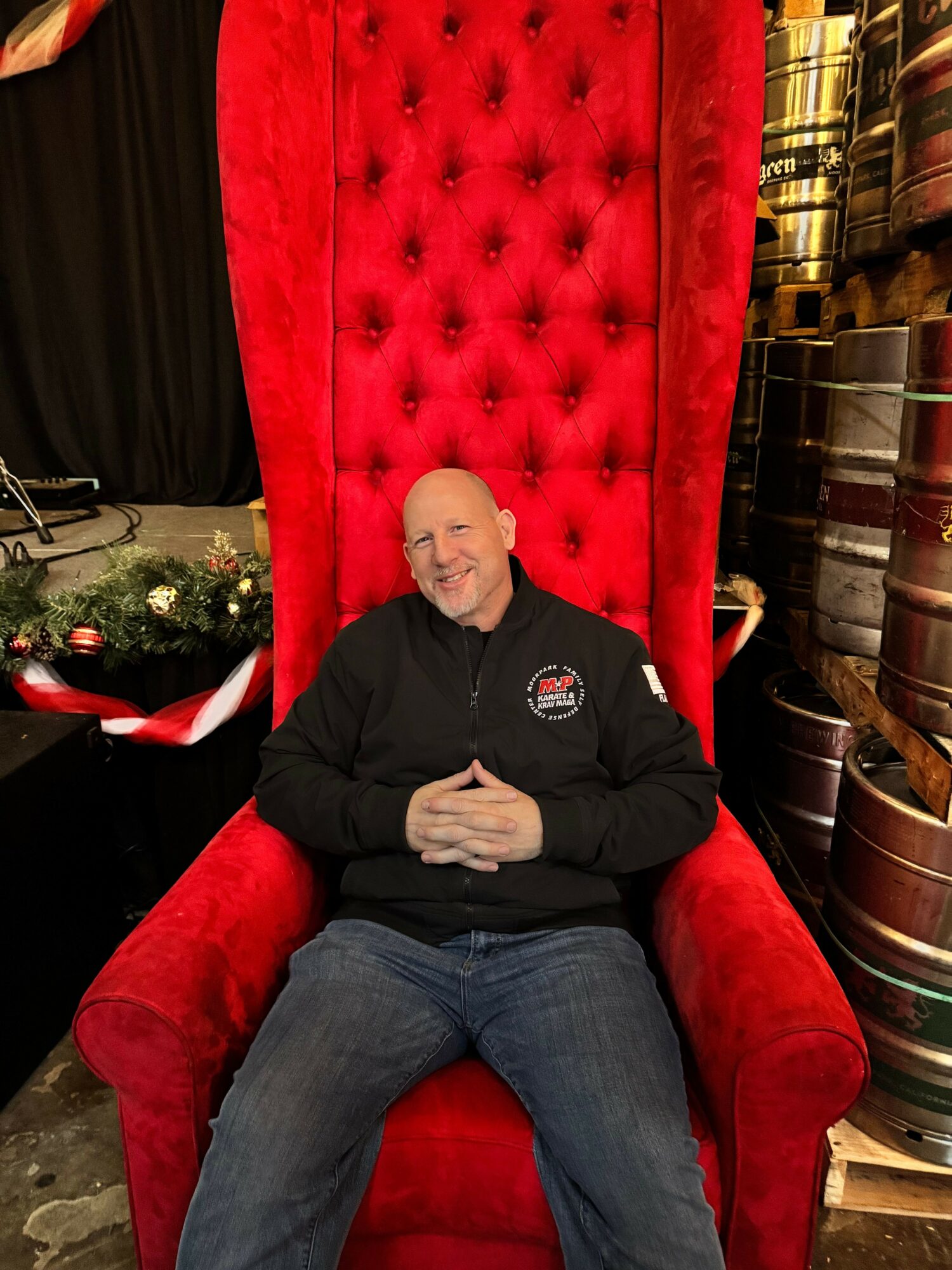Man sitting on a large red throne-like chair with a high back, smiling, in a decorated indoor setting.