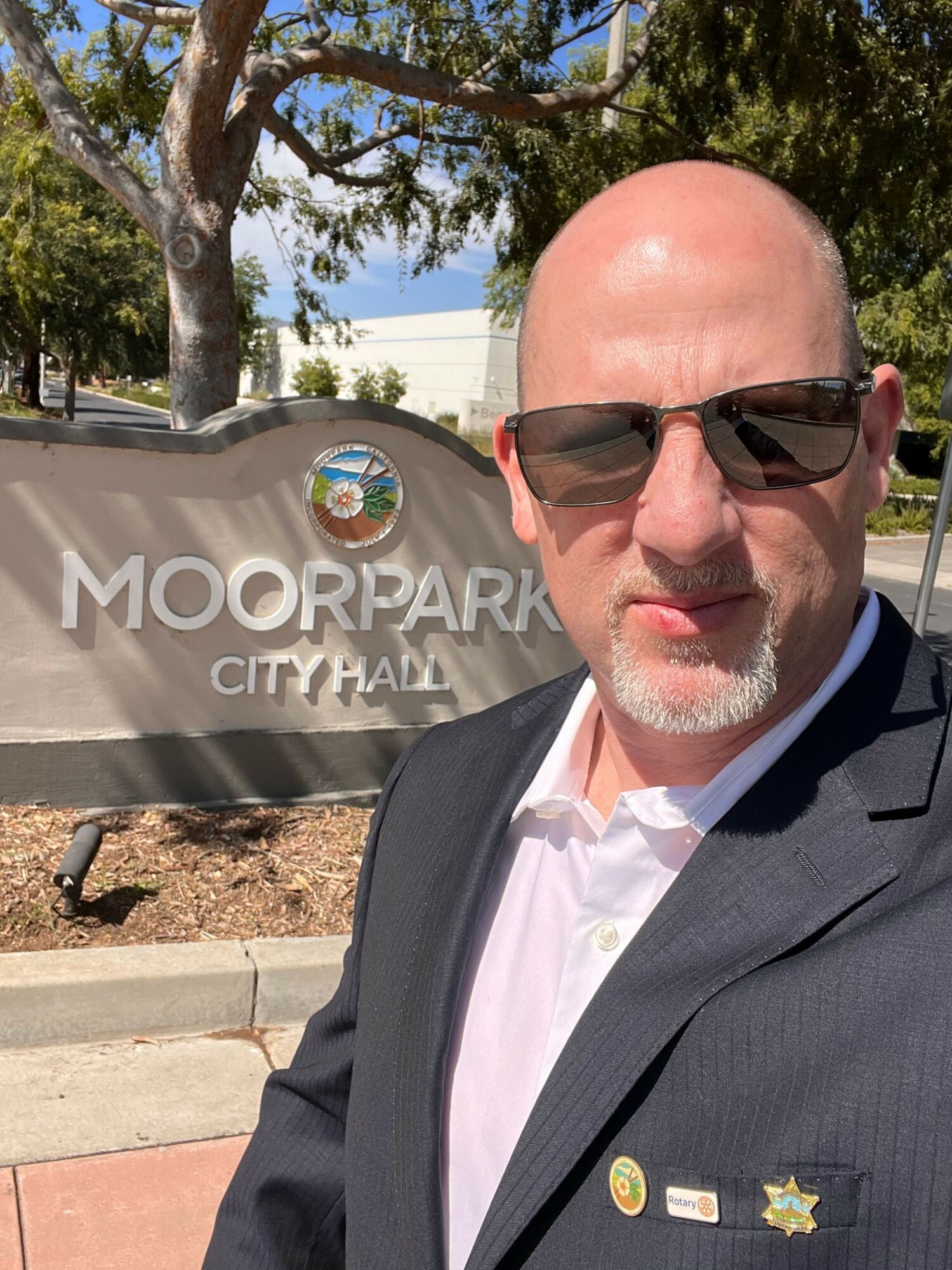 Man with glasses and a suit taking a selfie outdoors near a sign that reads 'Moorpark City Hall'.