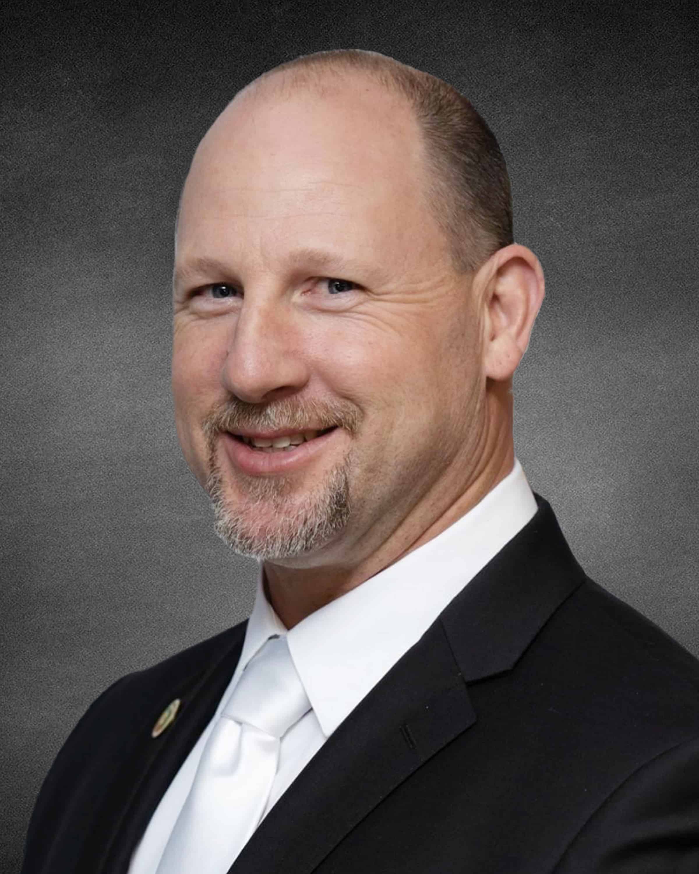 Man with short hair, beard, wearing a suit and white shirt, smiling against a dark background.