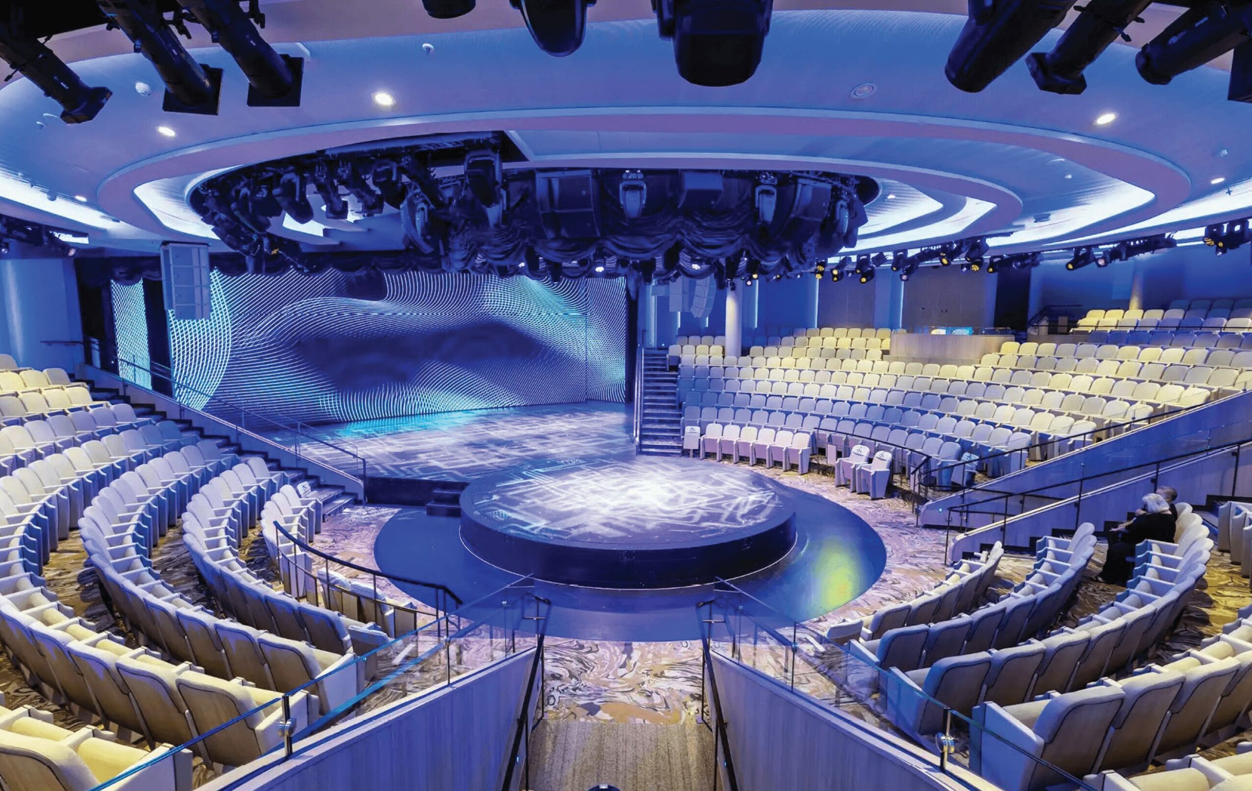Empty auditorium with a stage, curved seating, and blue lighting, viewed from the back center.