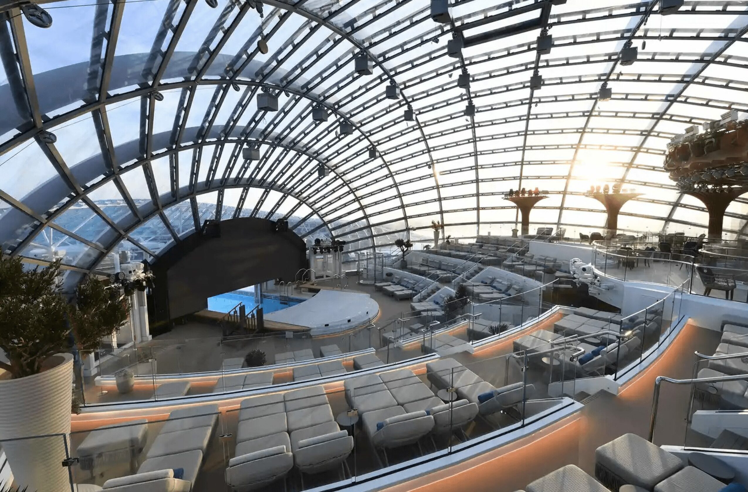 Indoor space with curved glass ceiling, seating area, and plants, illuminated by sunlight, with a view of the sky.