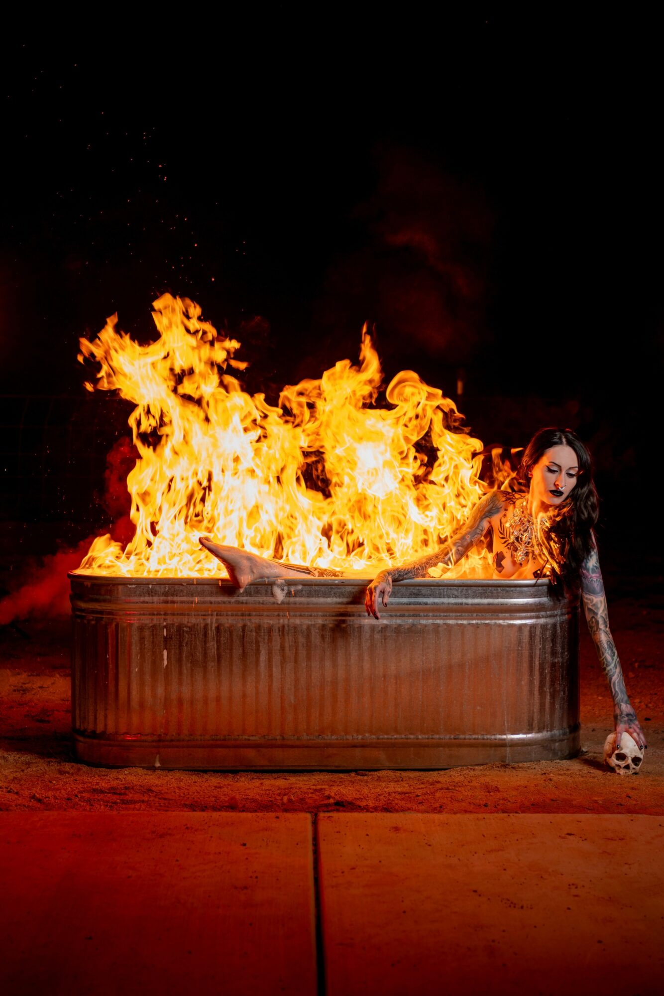 Person lying in a metal tub with flames behind them, holding a skull, at night.