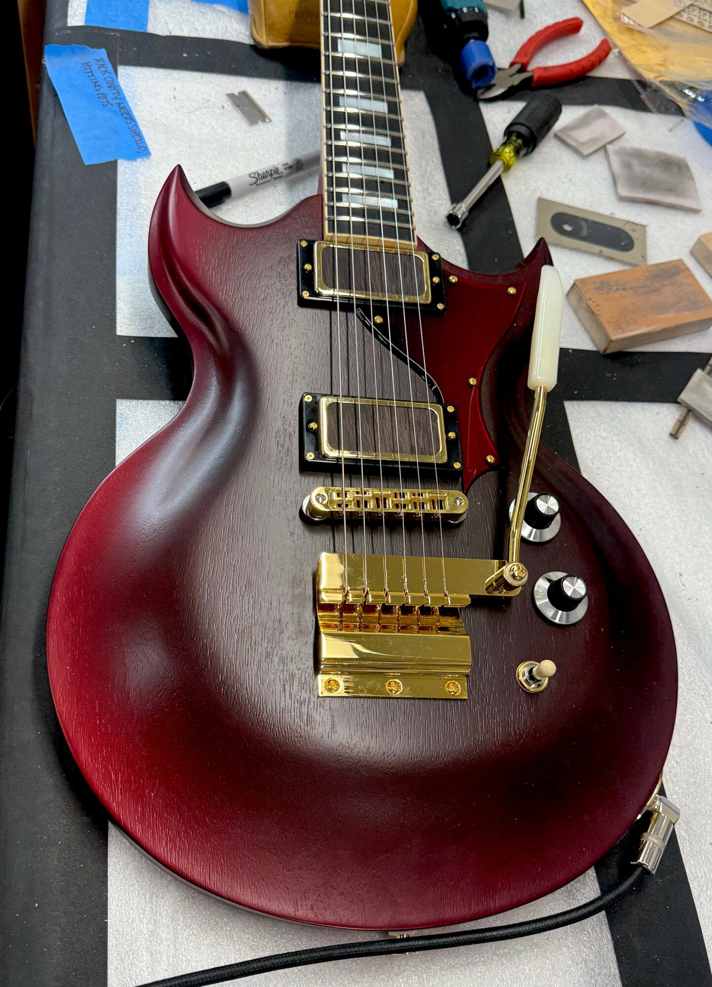 Red electric guitar with gold hardware and two pickups, on a workbench with tools and parts.