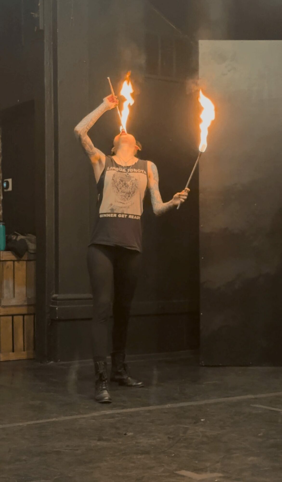 Person performing fire breathing with flames in each hand, standing indoors against dark walls.