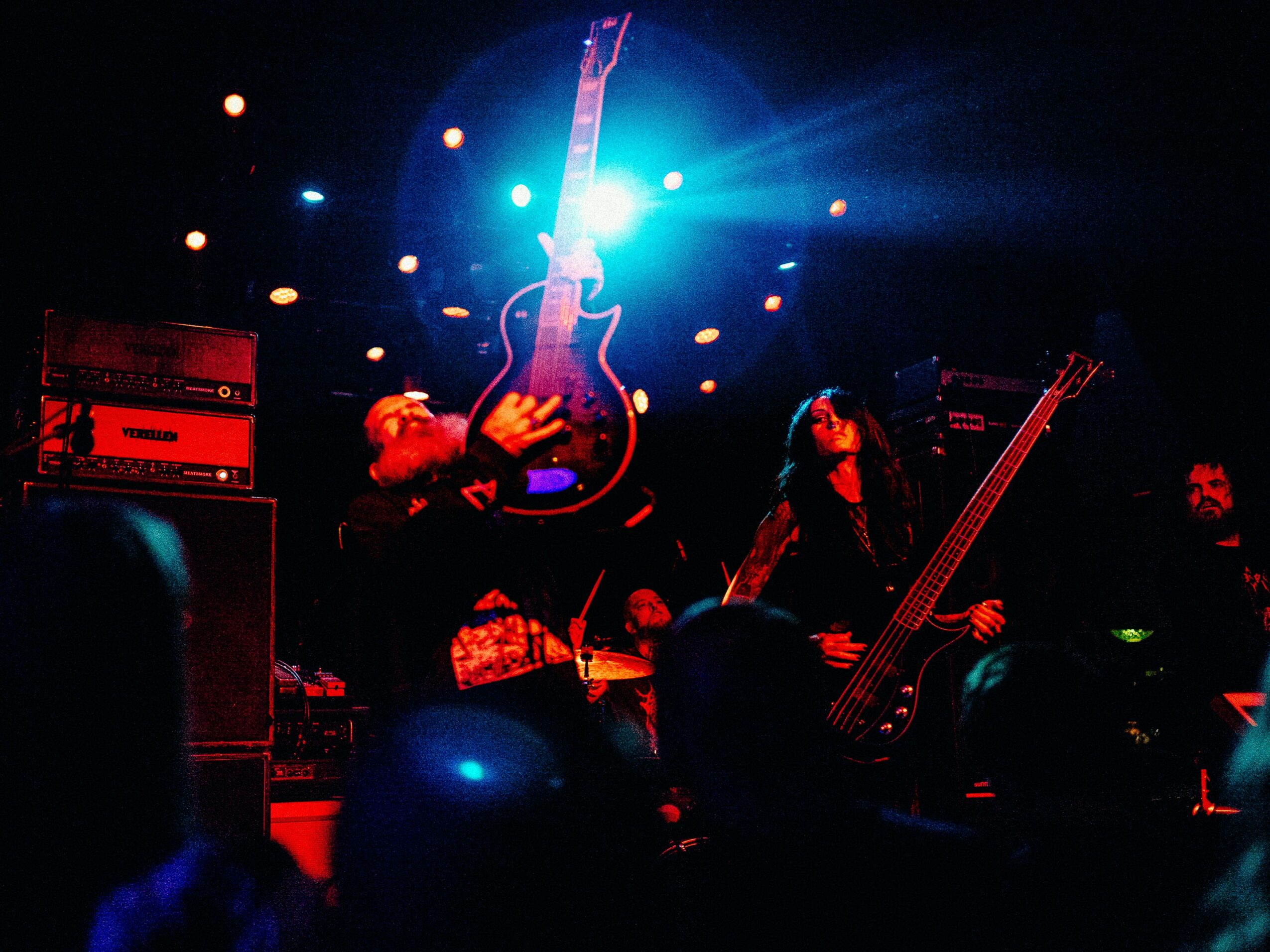 Musicians perform on stage with a guitar hanging above them, stage lights shining brightly, and equipment visible.