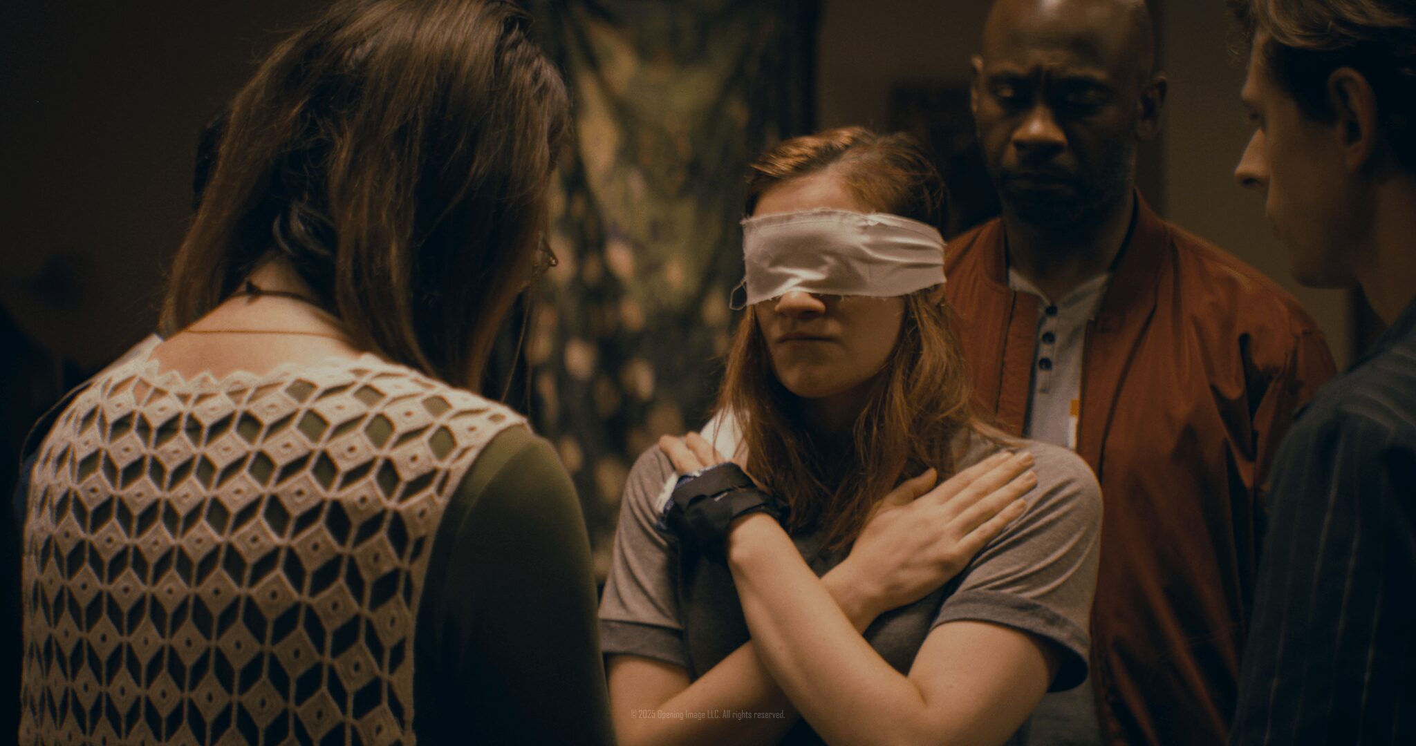 Four people in a dimly lit room, one woman with a blindfold, others observing her.