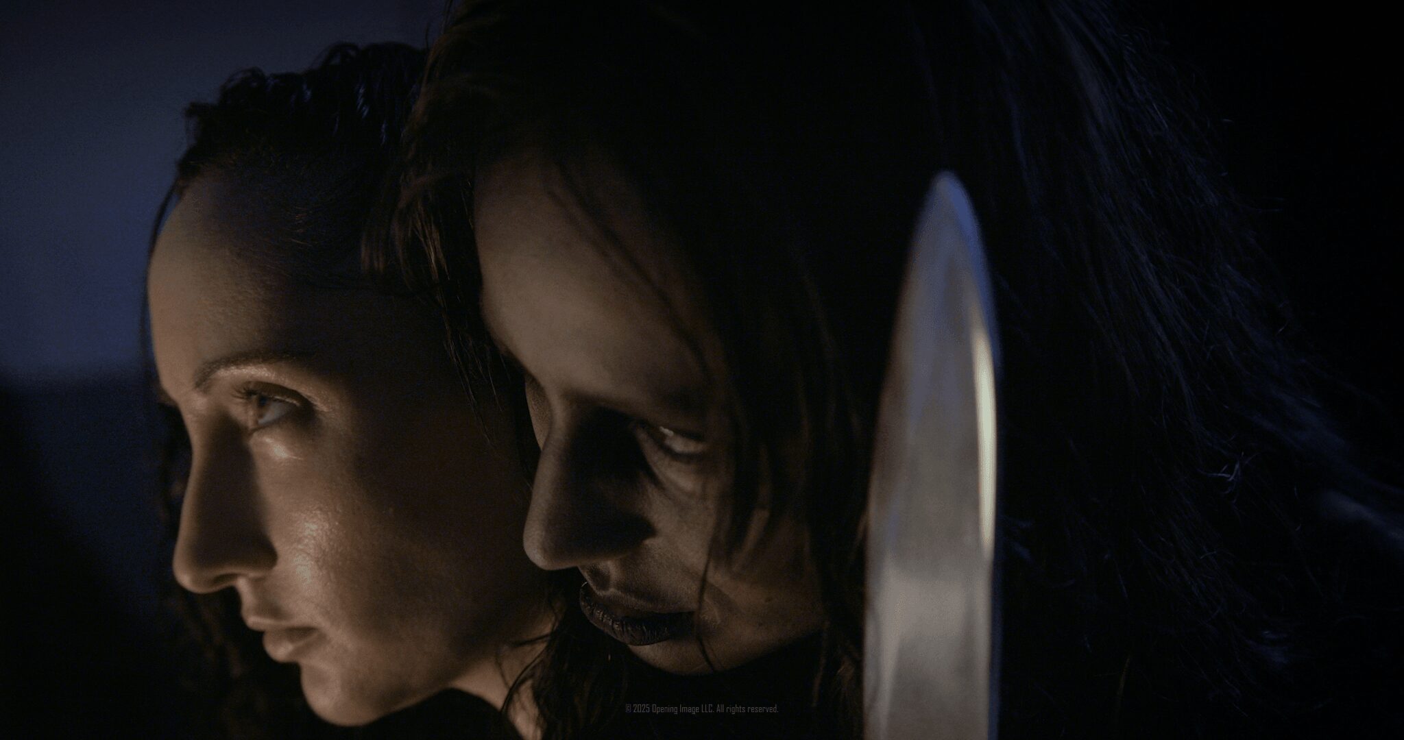 Two women with serious expressions, one with dark hair and the other with lighter hair, close together in low light.