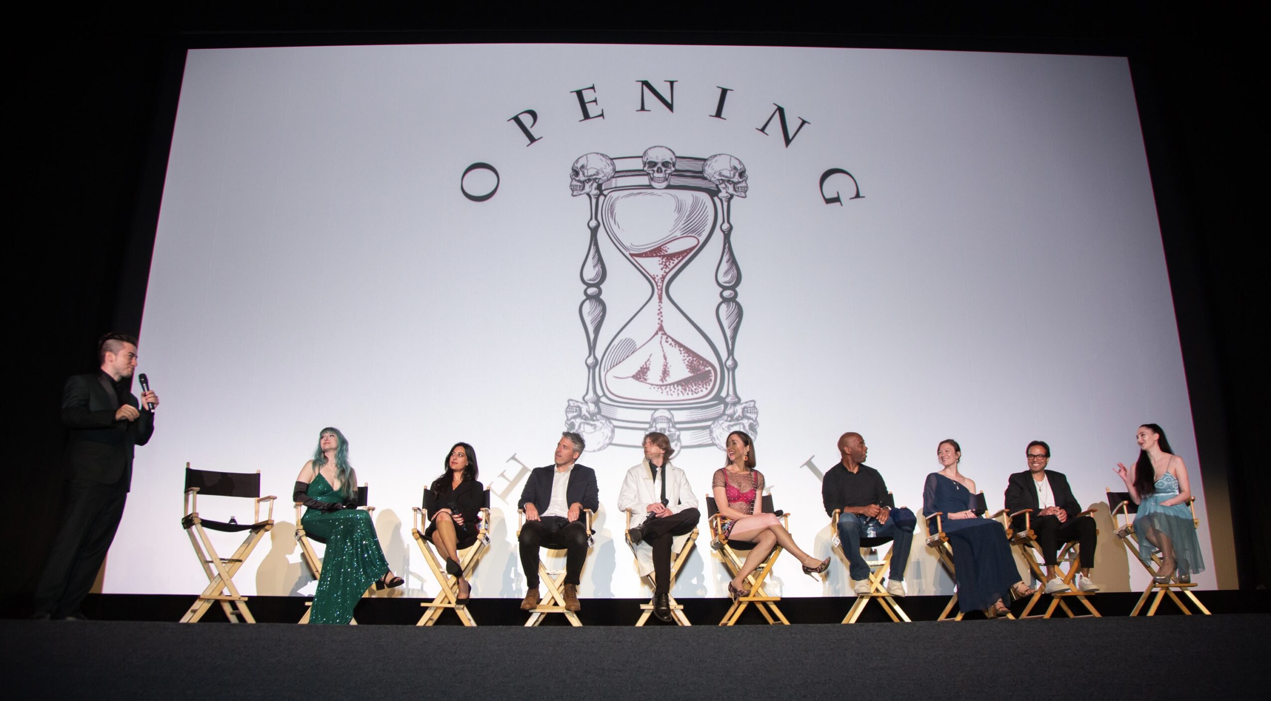 Panel of nine people seated on stage with a large screen behind them displaying an hourglass and the word 'Opening'.