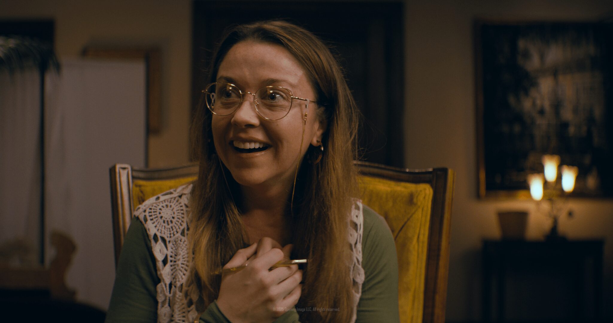Woman with glasses smiling, sitting at a table in a warmly lit room.