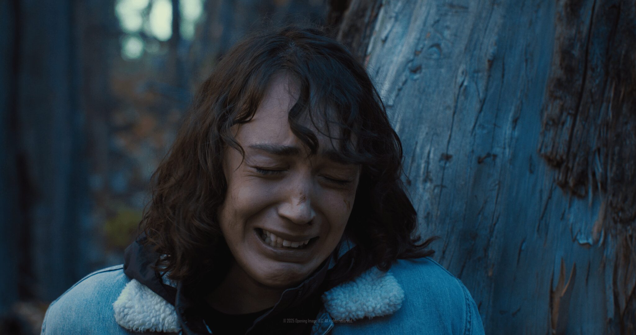 Person with curly hair crying outdoors near a wooden wall.