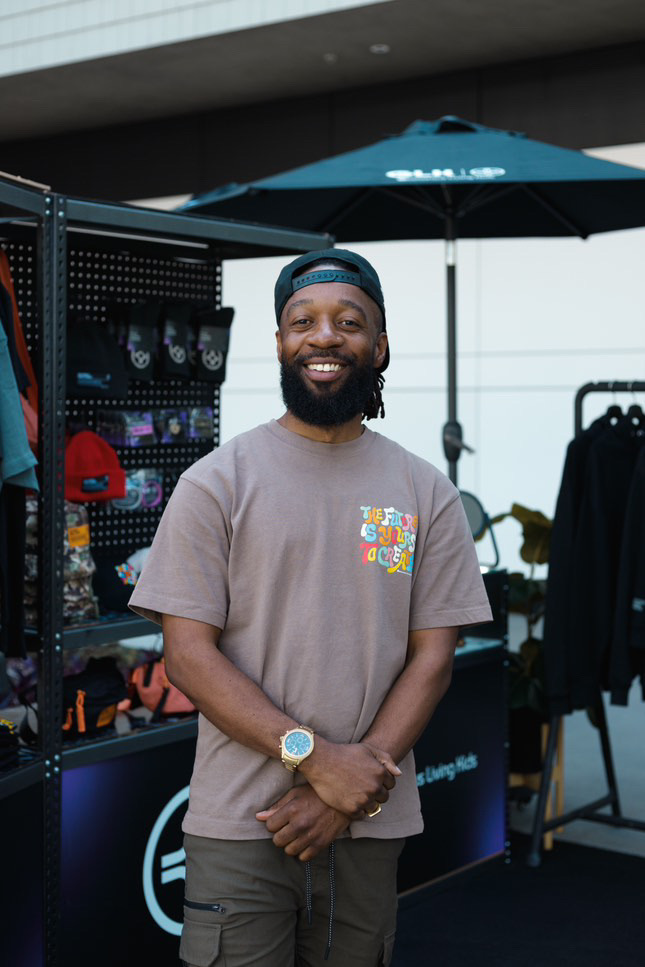Smiling man with beard, wearing a gray t-shirt, standing outdoors near a display with hats and accessories.
