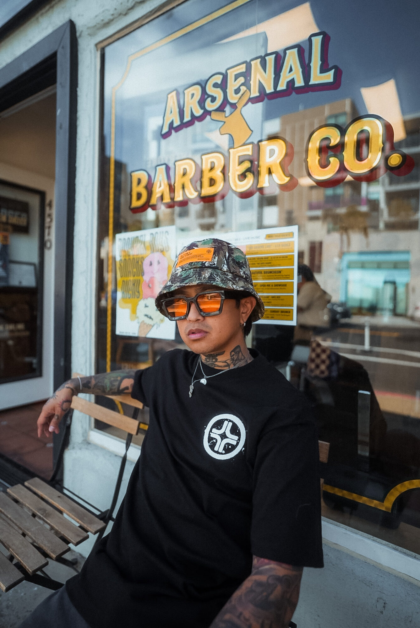 Person with tattoos wearing sunglasses and a bucket hat sitting outside barber shop with sign reading Arsenal Barber Co.