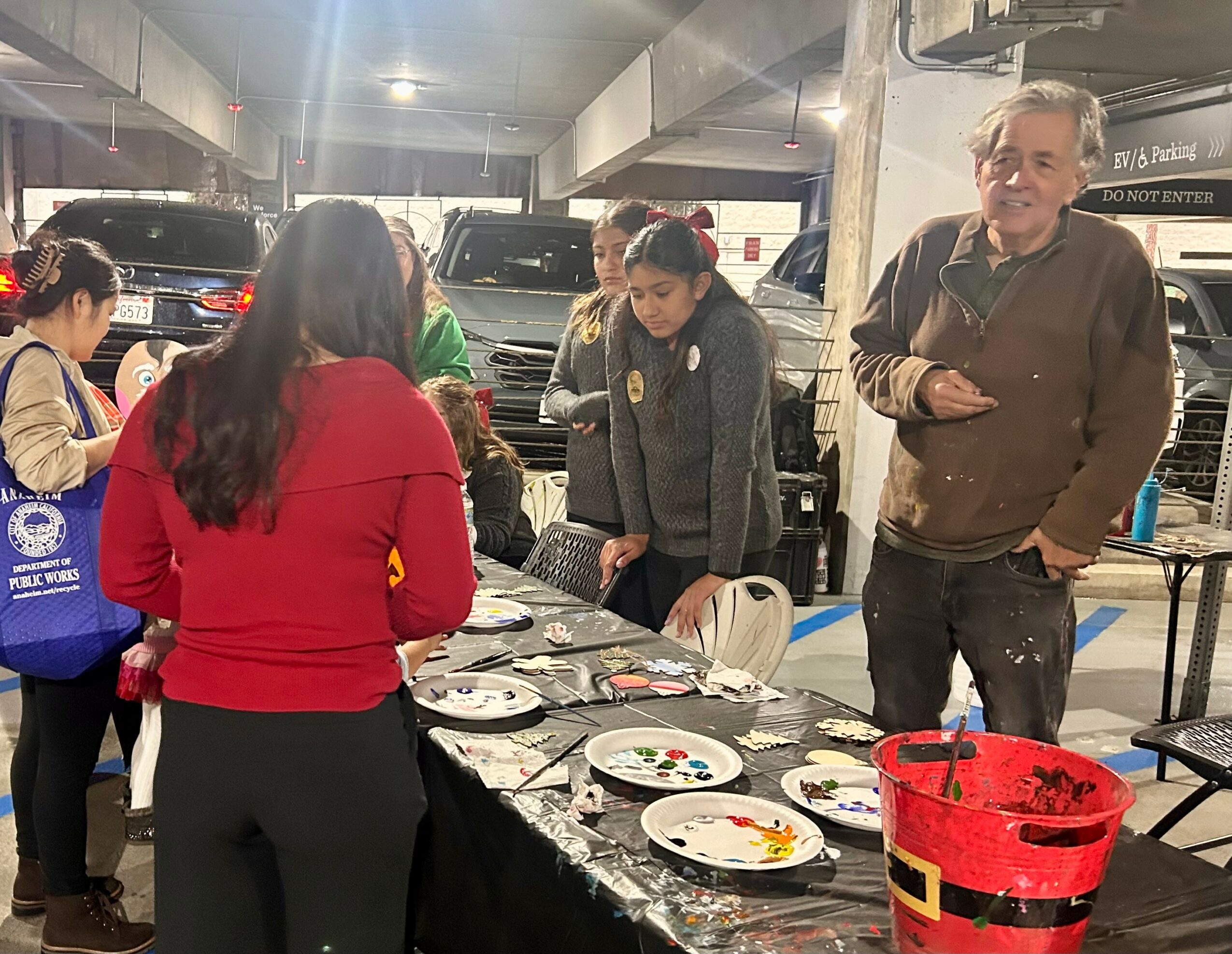 Group of people gathered around a table with plates and a red bucket in a parking garage.