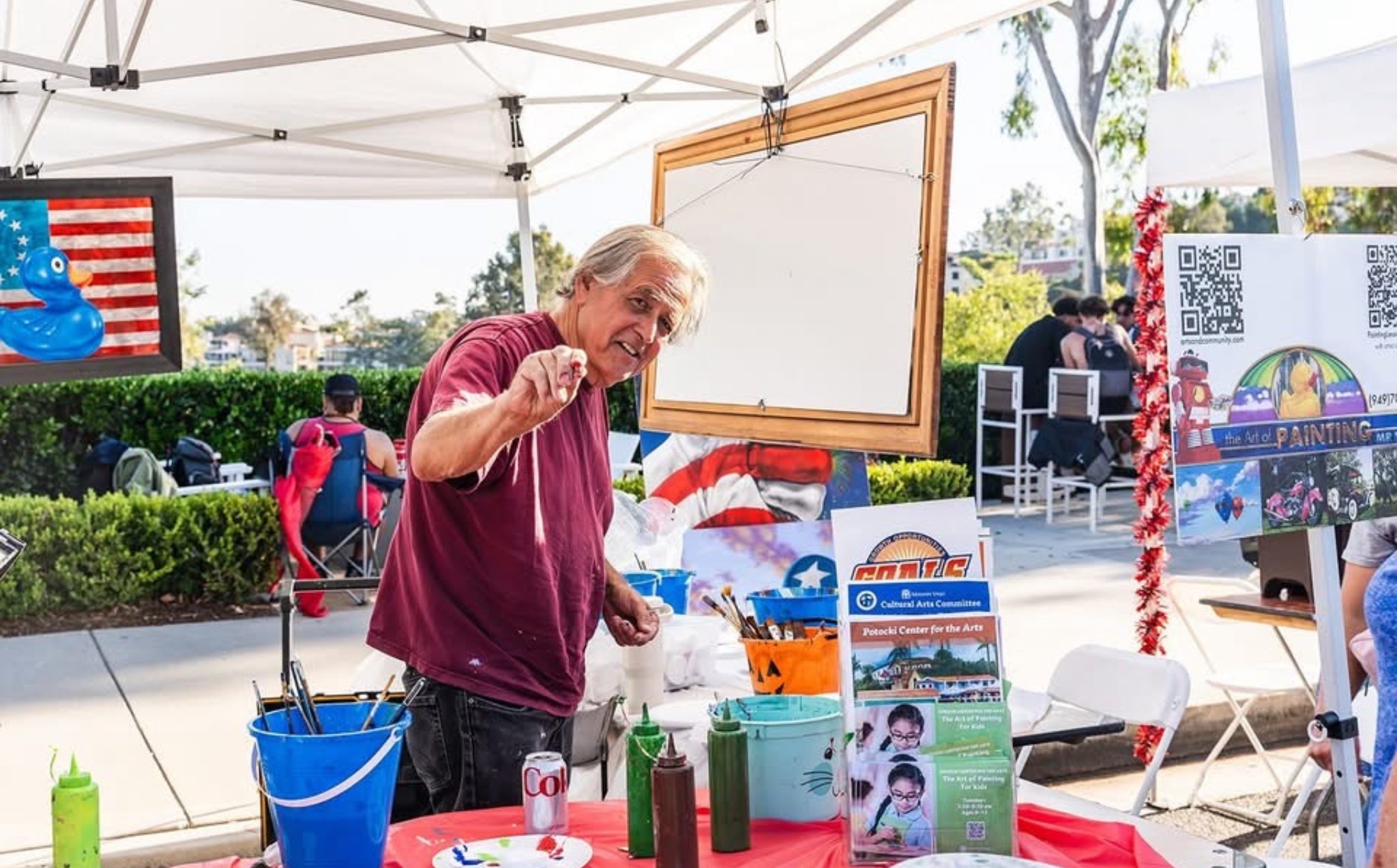 Elderly man smiling and waving at outdoor event under a canopy with tables and promotional materials.