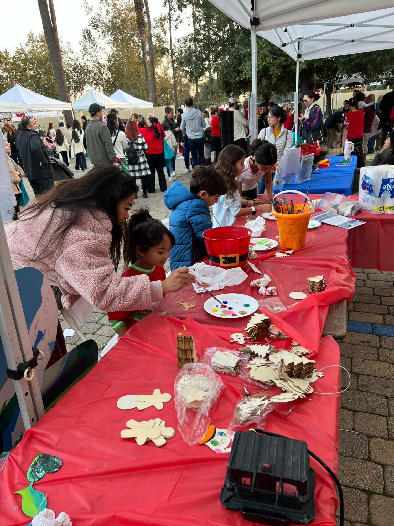 People gather outdoors around tables with crafts and snacks under tents, trees in background, sunny day.