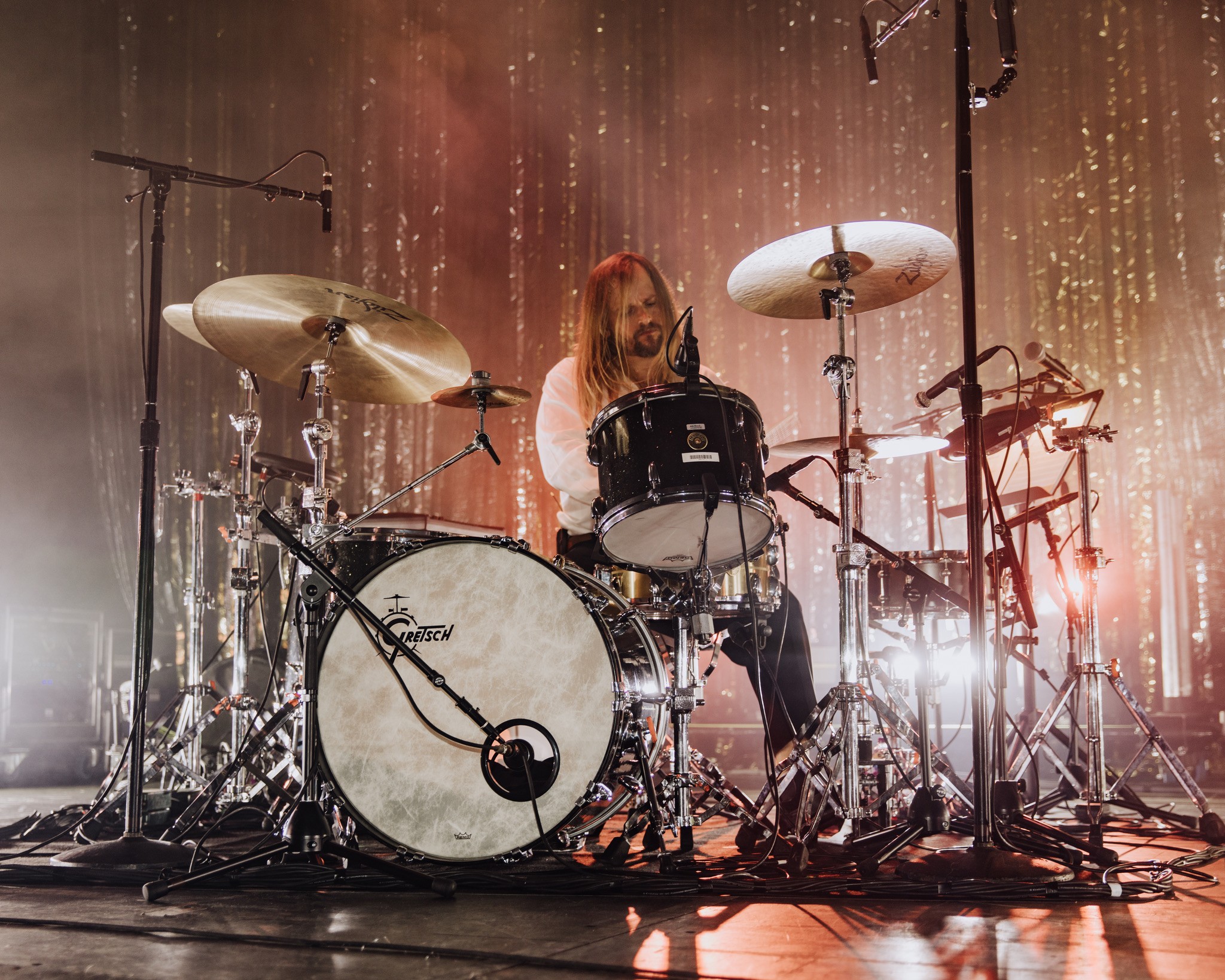 Person playing drum set on stage with golden curtain backdrop and stage lighting.