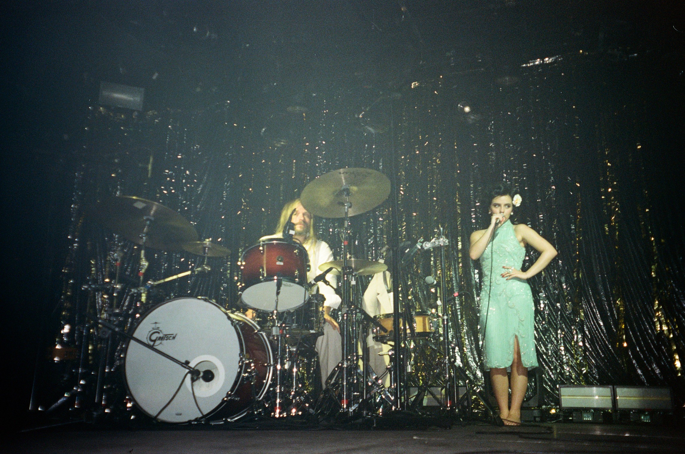 Two women on stage with a drum set and shiny curtain backdrop, one singing and the other playing drums.