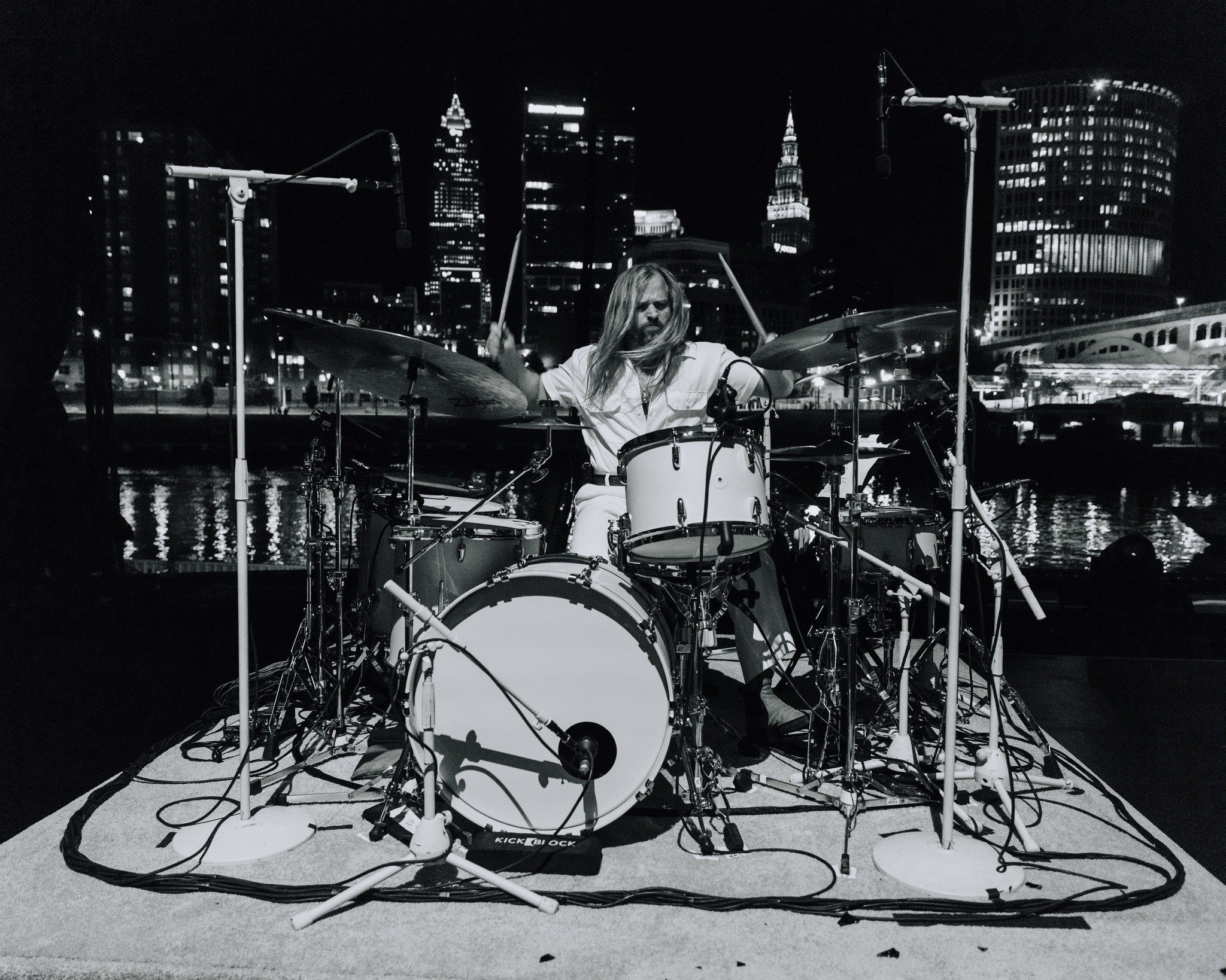 Person playing drums on stage with city skyline in background at night.