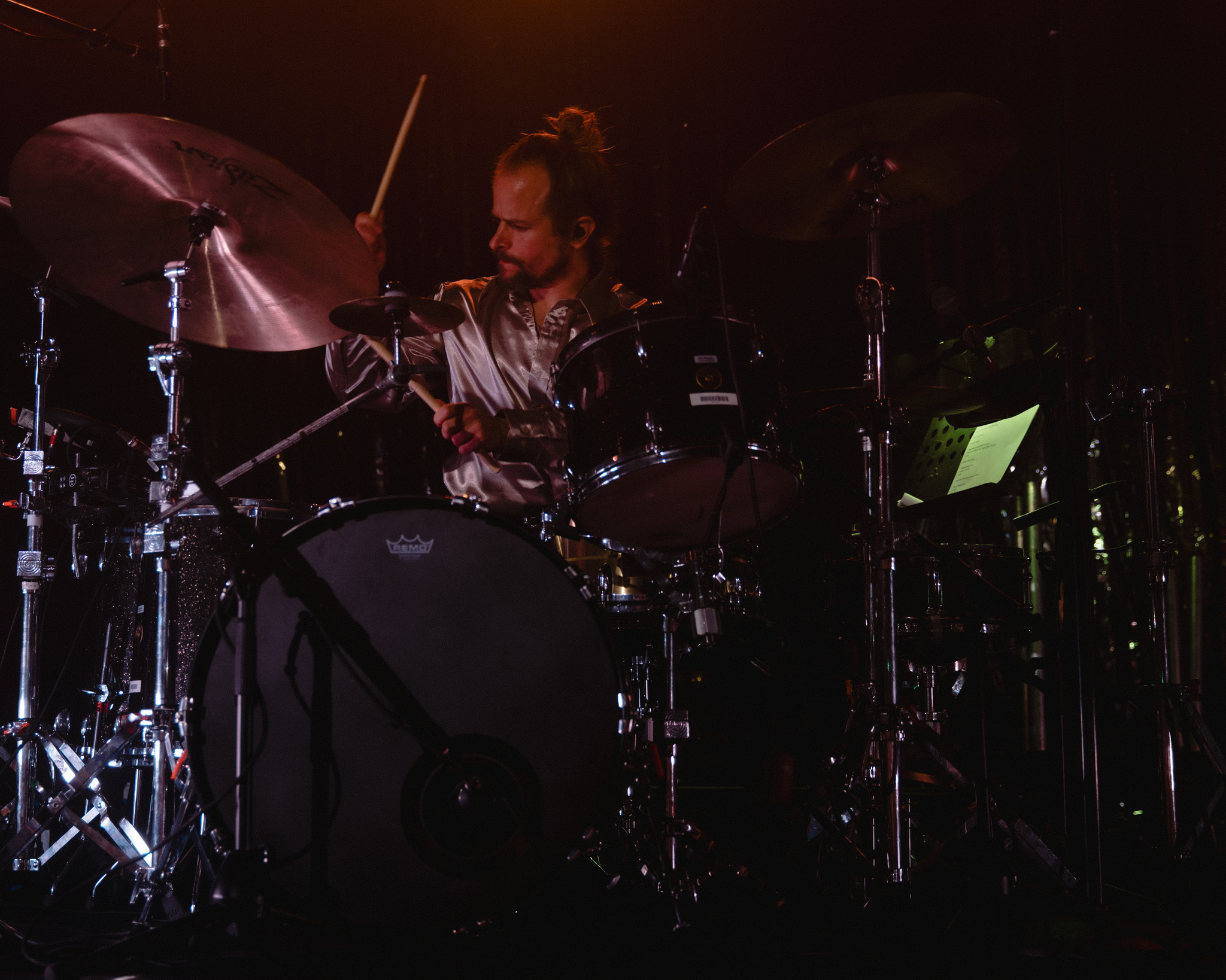 Person playing drums on stage with cymbals and drum set, dark background, stage lighting.