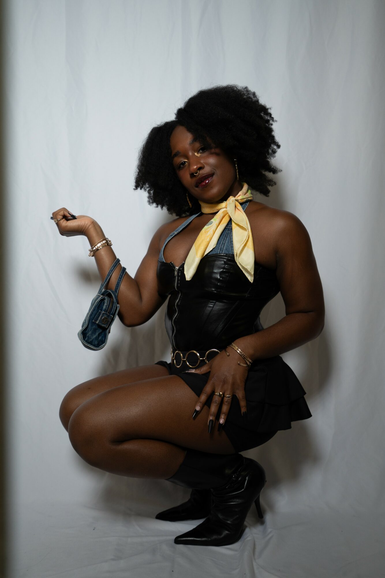 Woman with curly hair in black outfit and yellow scarf, posing with one knee up, holding a small bag, against a white backdrop.
