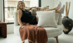 Woman sitting on a white sofa with pillows, in a bright room with large window and modern decor.