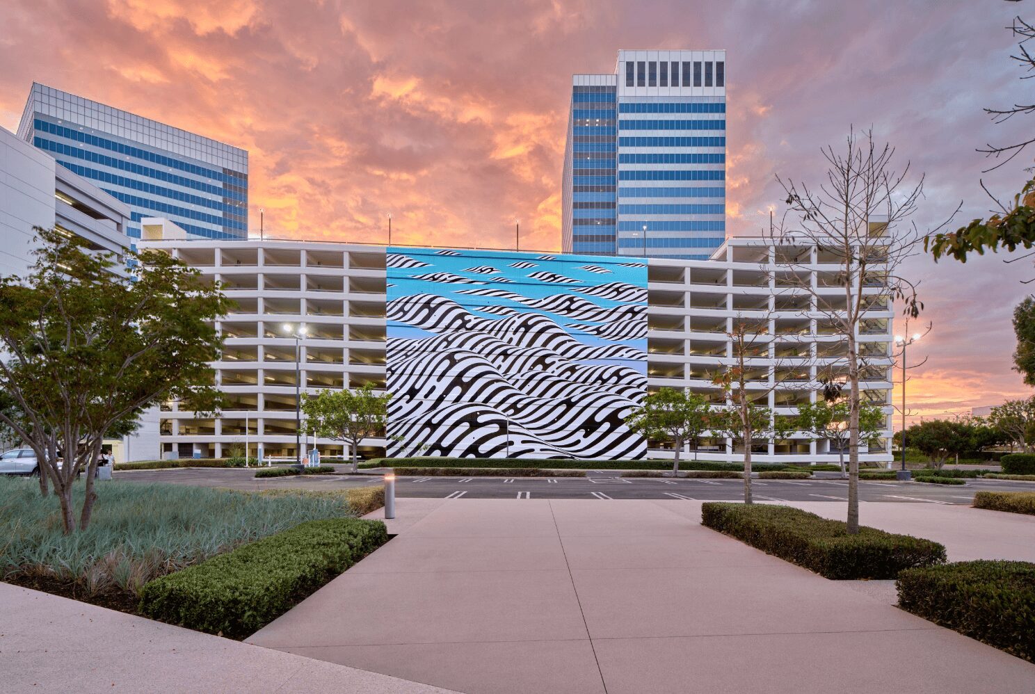 Modern building with a large patterned mural, surrounded by trees and walkways, under a colorful sunset sky.