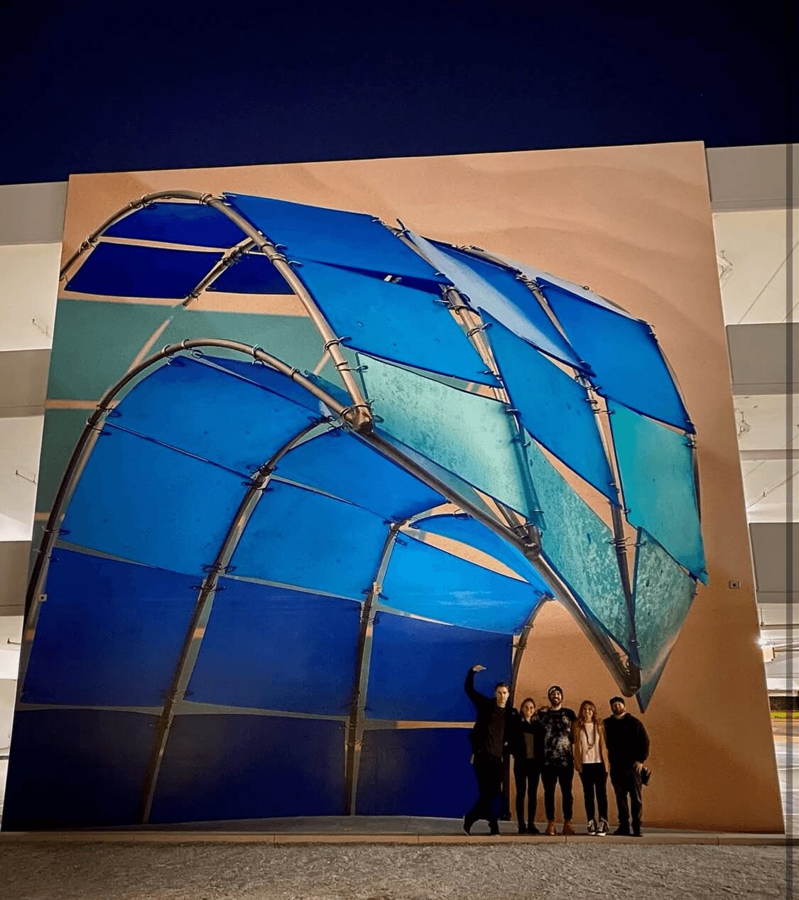 Group of people standing in front of a large, abstract blue and green sculpture at night.