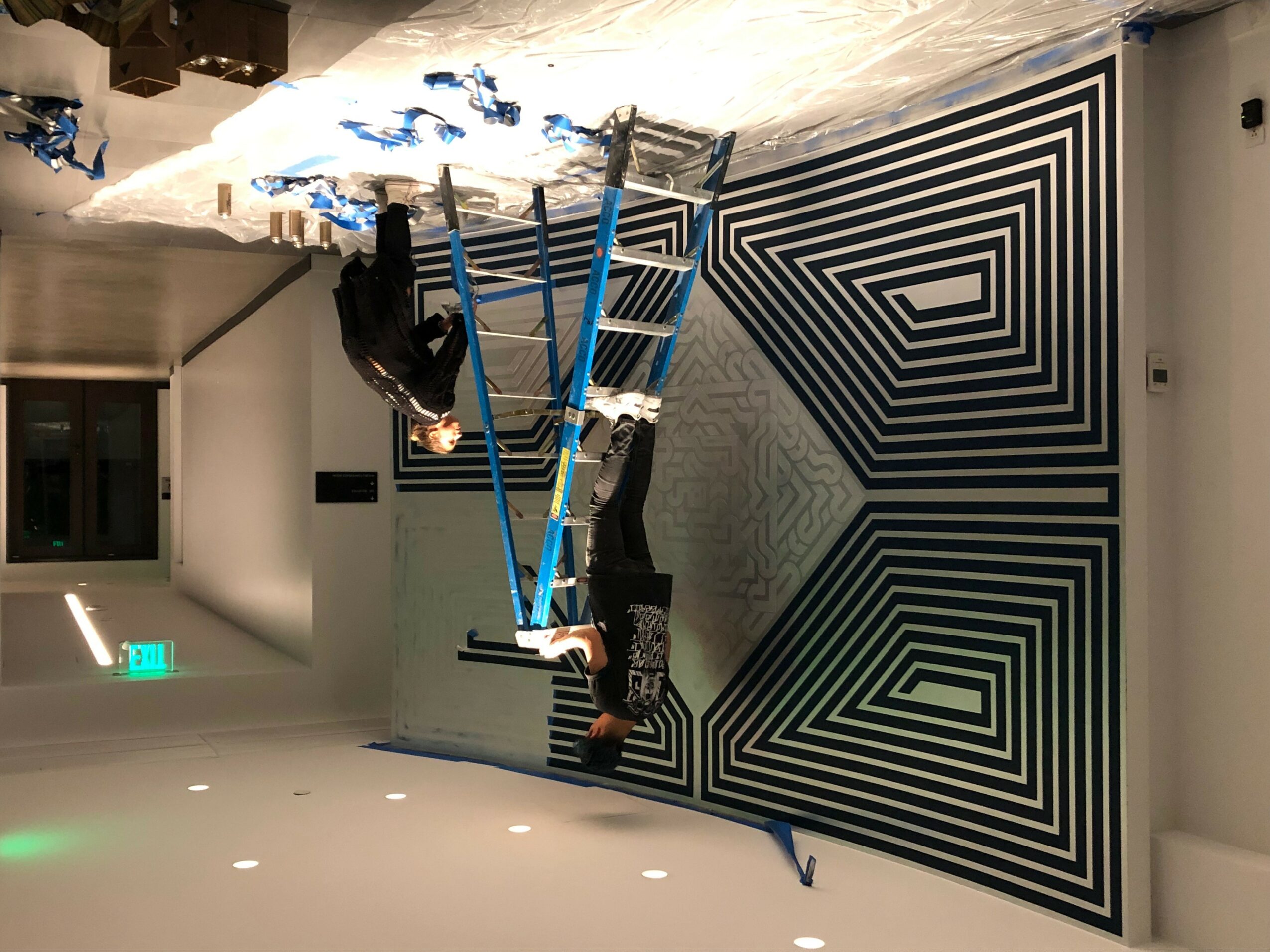 Two people are on a blue and white climbing structure in front of a black and white geometric wall pattern.