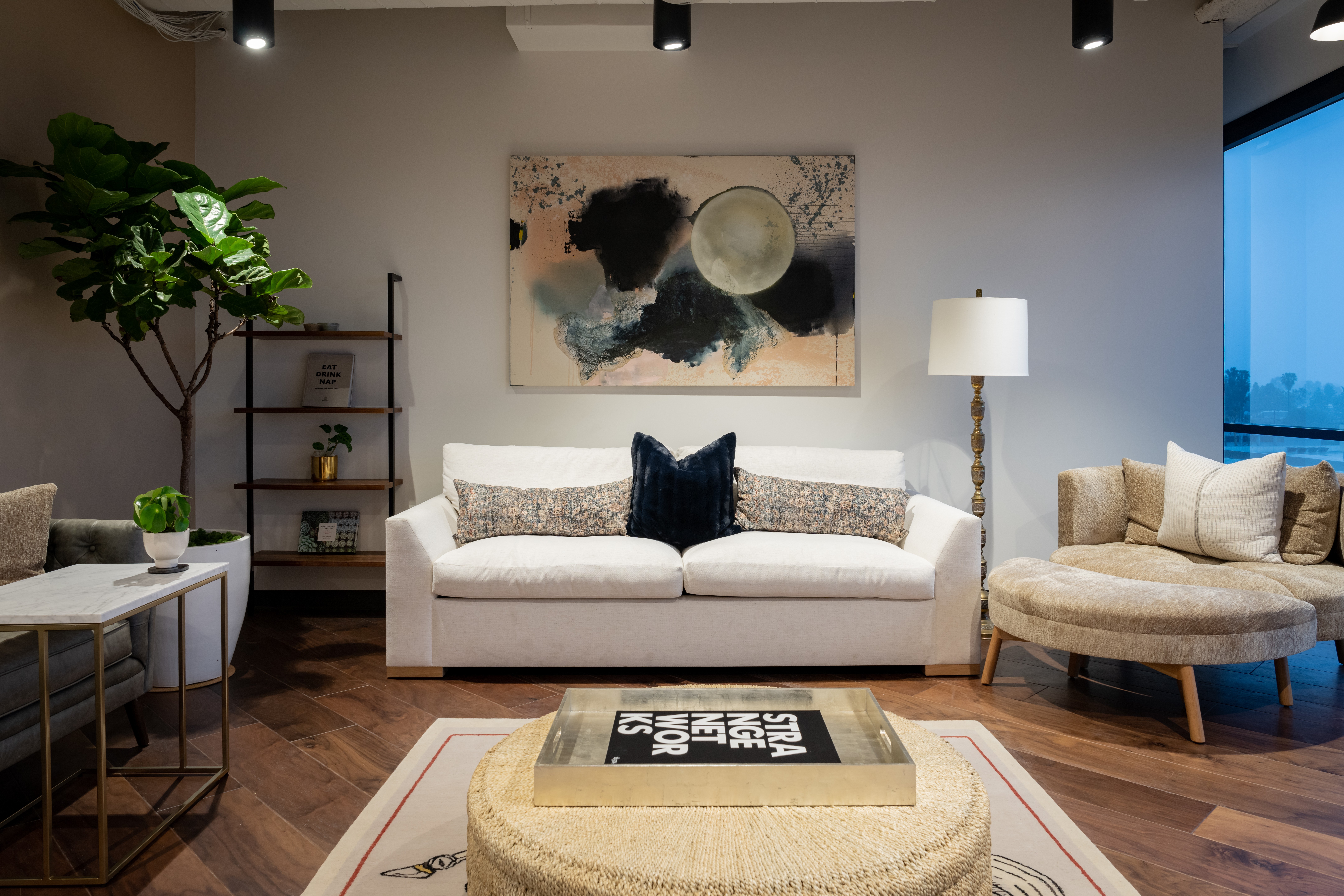 Living room with white sofa, beige armchair, tall lamp, artwork, and large window with blue sky view.